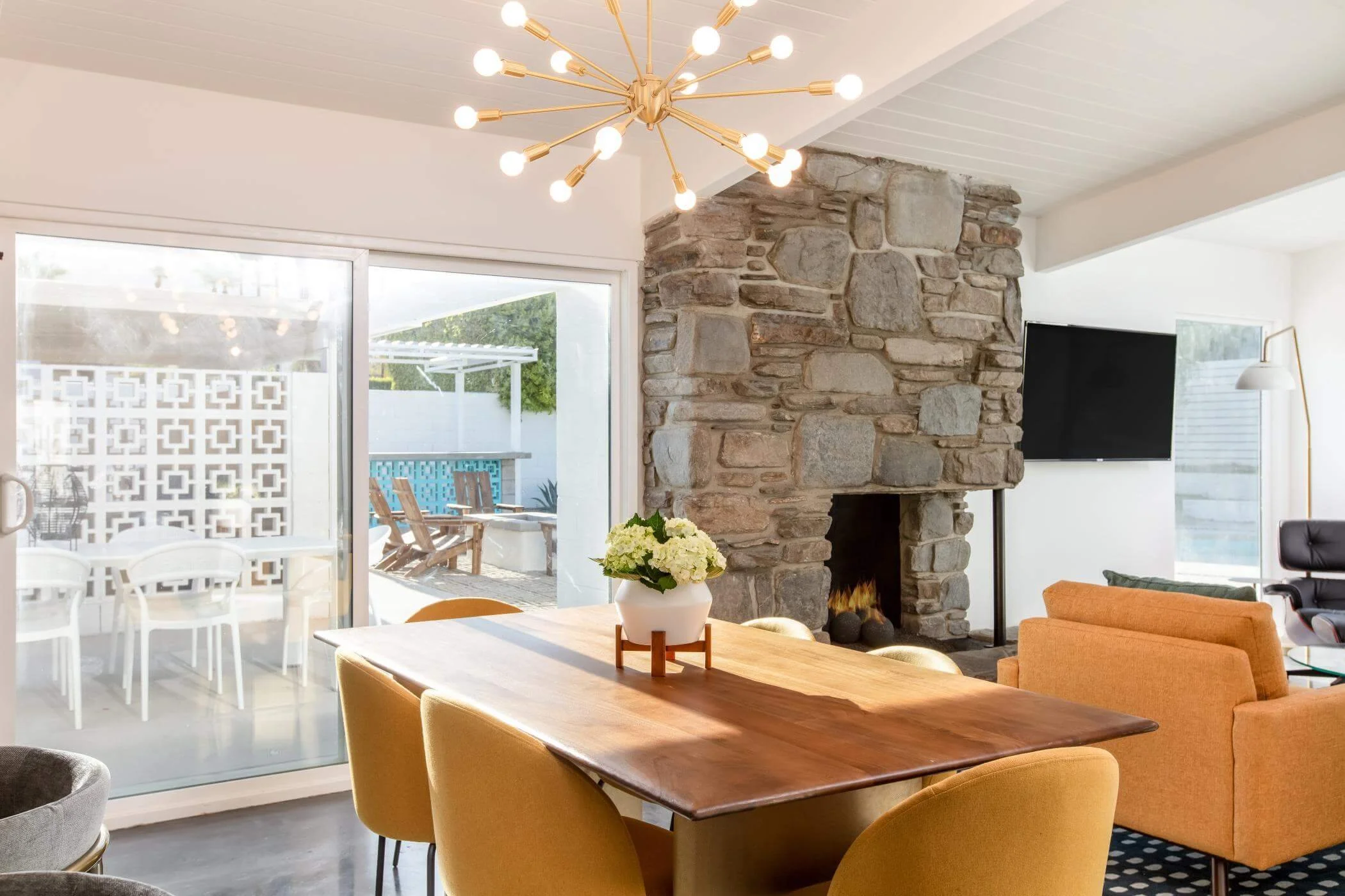Living room with a stone fireplace, a wooden dining table with yellow chairs, orange sofas, a wall-mounted TV, large glass sliding door leading to outdoor patio with outdoor furniture, modern chandelier overhead, and natural light streaming in.