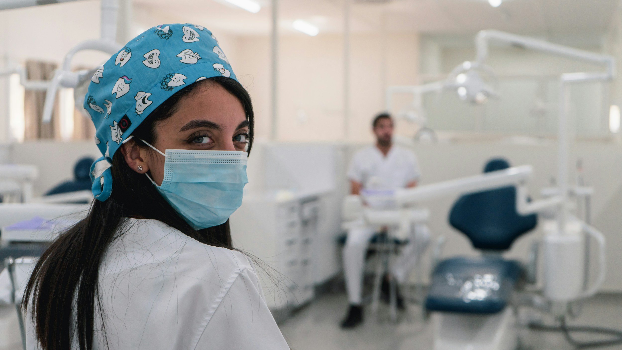 A female healthcare worker wearing a blue surgical cap with white cartoon faces and a blue face mask, looking over her shoulder in a medical setting with dental chairs and a male healthcare worker in the background.