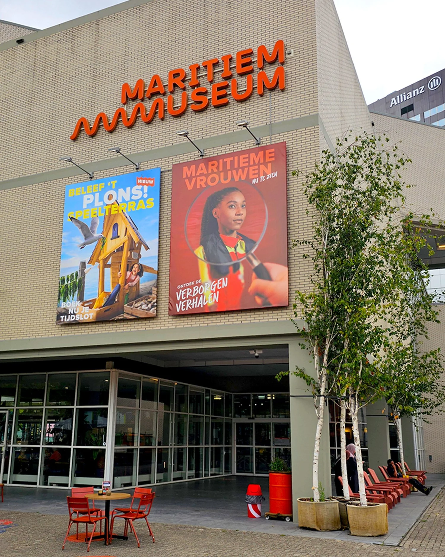 Exterior view of the Maritime Museum building with large red signage and colorful banners, outdoor seating area with orange chairs and tables, trees, and a modern glass entrance.