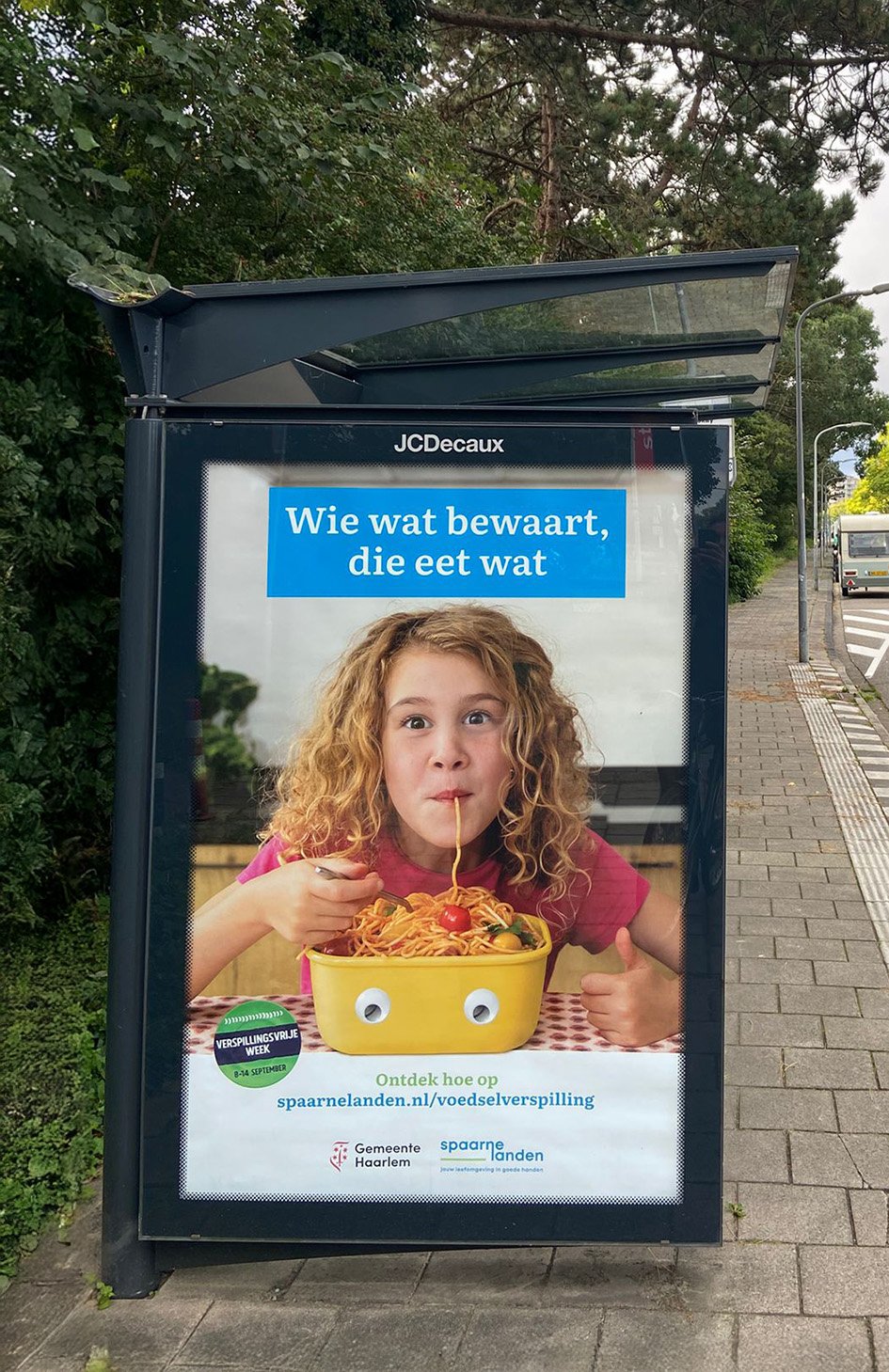 Bus stop advertisement showing a girl eating spaghetti, with Dutch text about conservation and food wastage, and logos of local organizations.