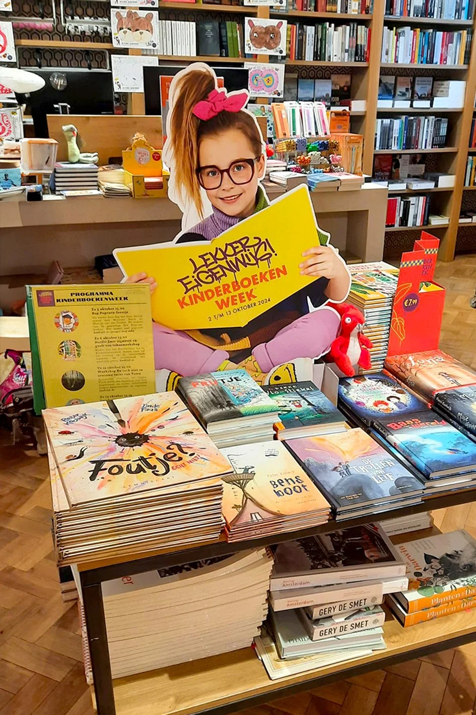 A display table with children's books and a cardboard cutout of a young girl holding a yellow sign promoting Kinderboekenweek (Children's Book Week). The table features various colorful books including titles like "Fout!" and "Ben's Boot". In the bac