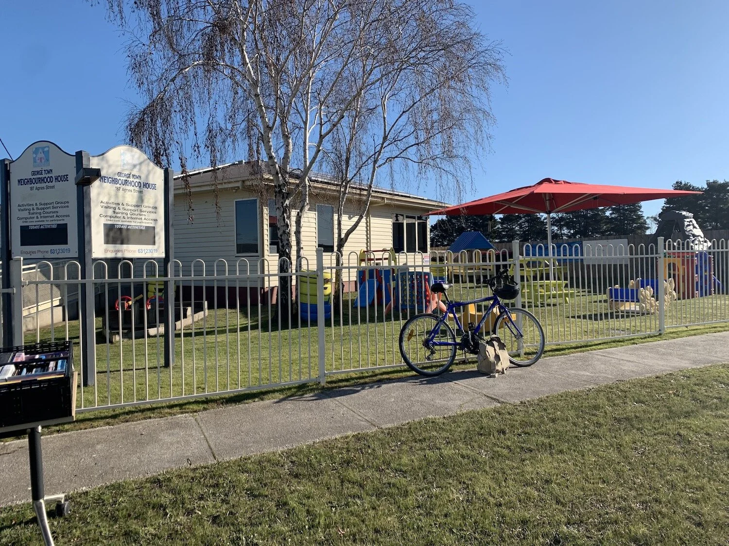 The outside of the george Town Neighbourhood House has childrens play equipment a big sunshade umbrella, a cream fence with a persons bicycle leaning against it