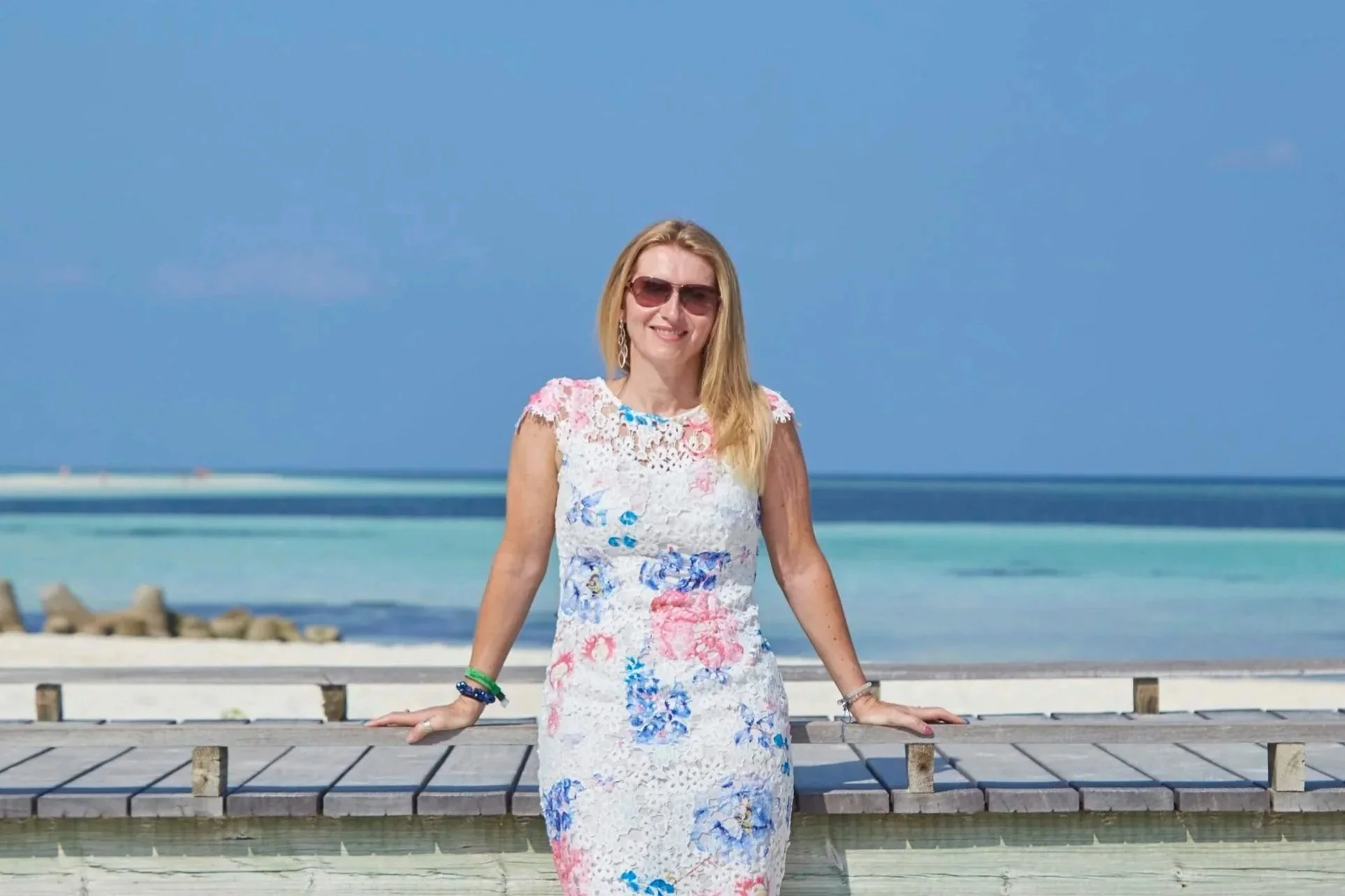 Woman in floral dress with sunglasses standing on a wooden pier in front of the ocean under a clear blue sky.