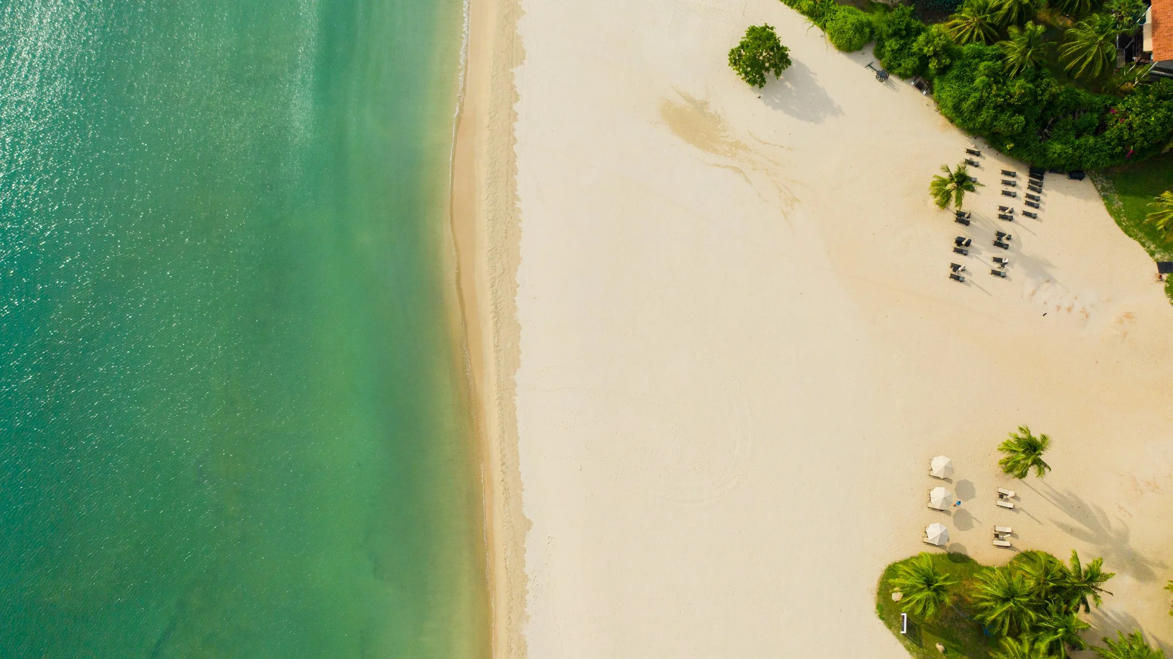 Aerial view of a tropical beach with turquoise water, white sandy shore, green trees, and outdoor seating under umbrellas.
