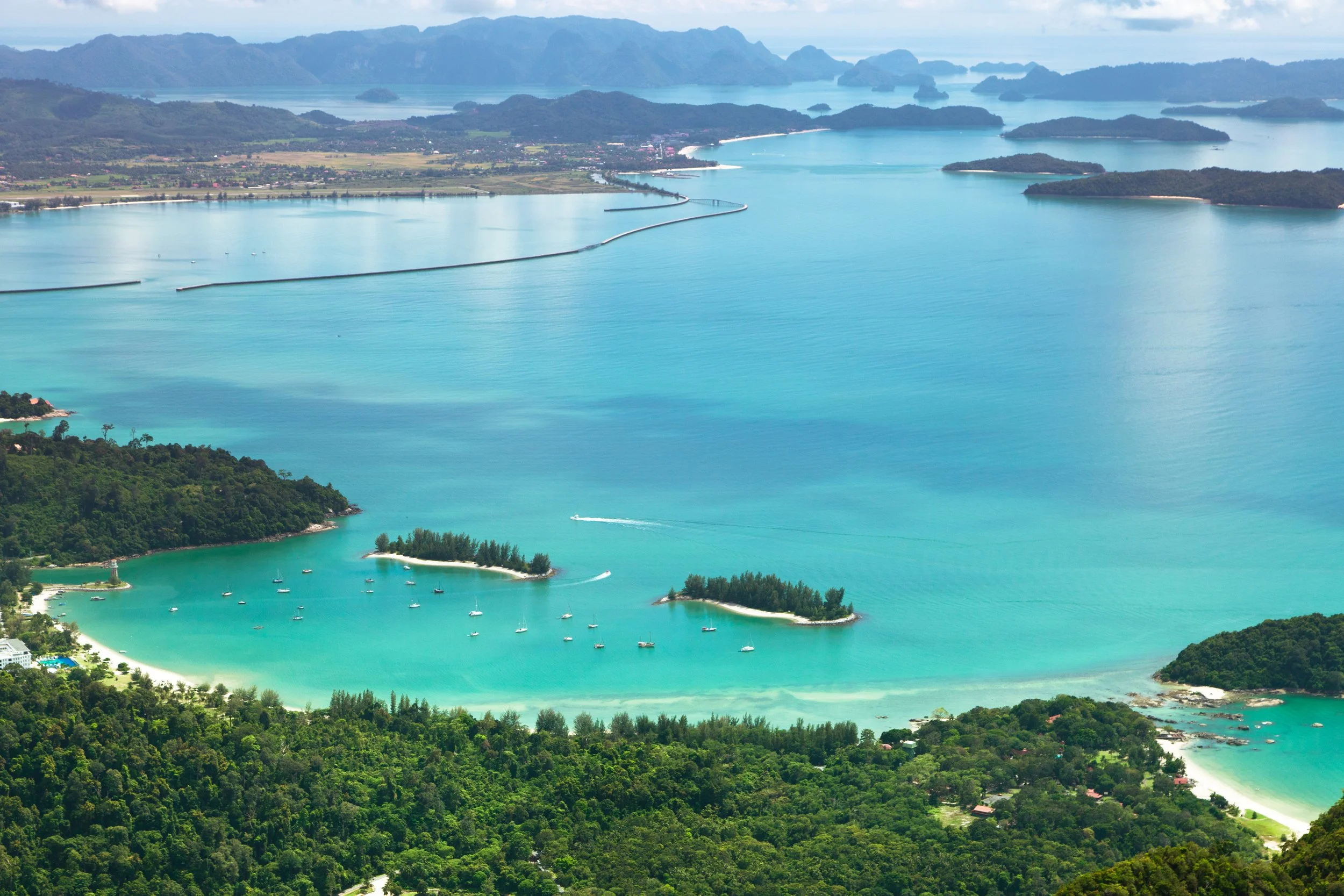 An aerial view of a turquoise coastal area with numerous sailboats anchored in the water, surrounded by lush green forests, hills, and distant mountains under a partly cloudy sky.