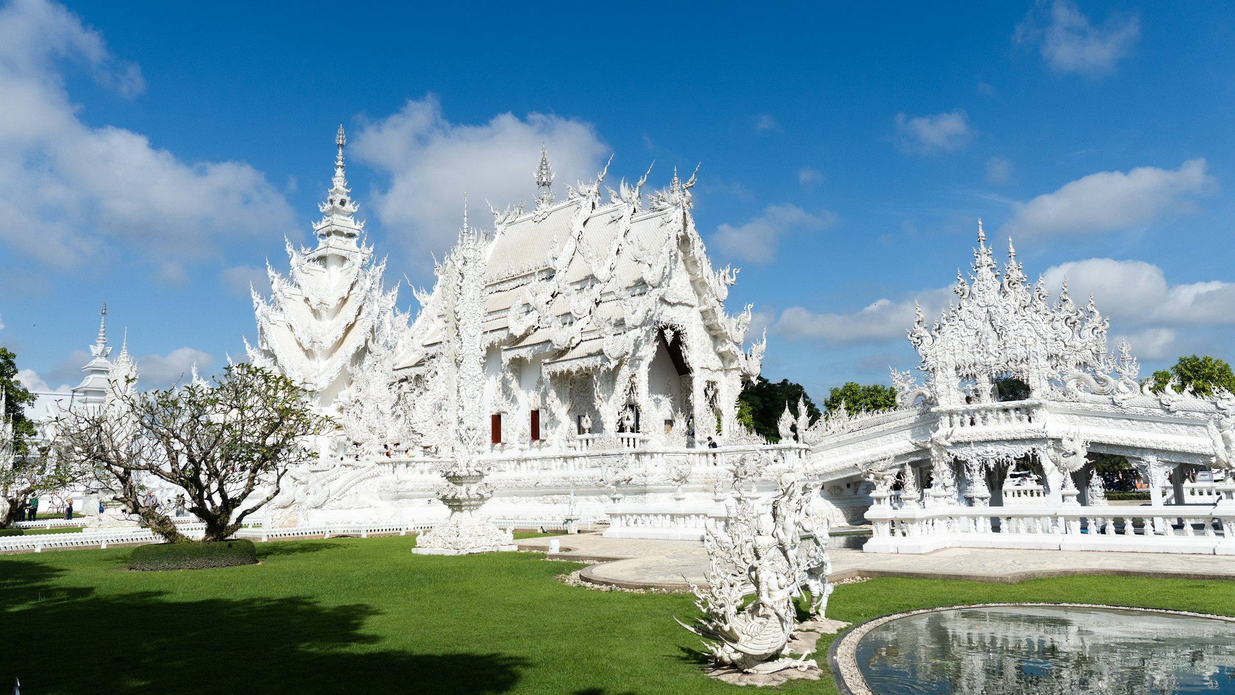 White ornate temple with intricate carvings, surrounded by greenery and a reflecting pool, under a blue sky with clouds.