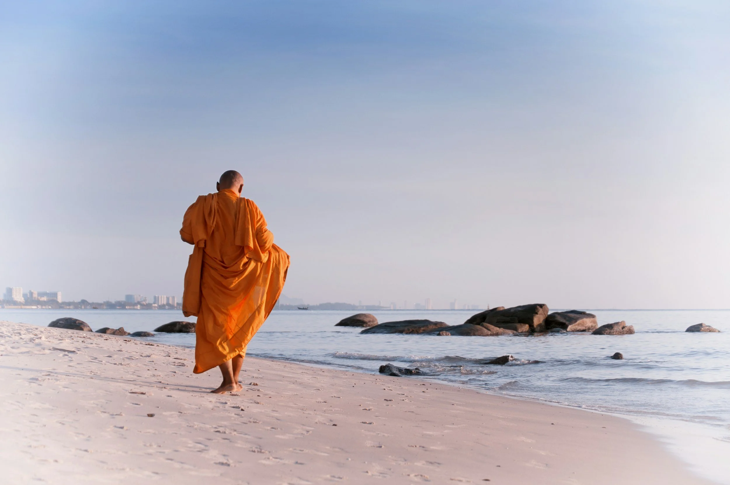 A Buddhist monk in orange robes walking on a sandy beach with rocks, looking towards the ocean and distant city skyline under a clear sky.