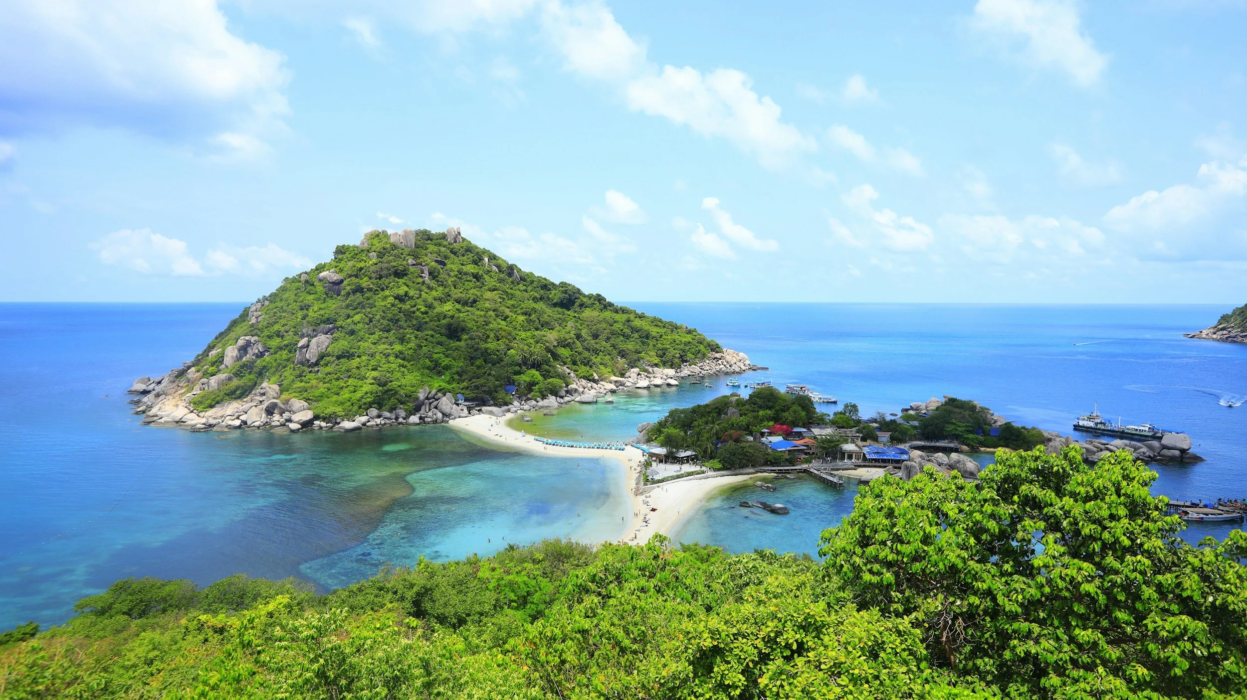 A lush green island with rocky hills, surrounded by turquoise waters, with boats docked near the shore and a small seaside village.
