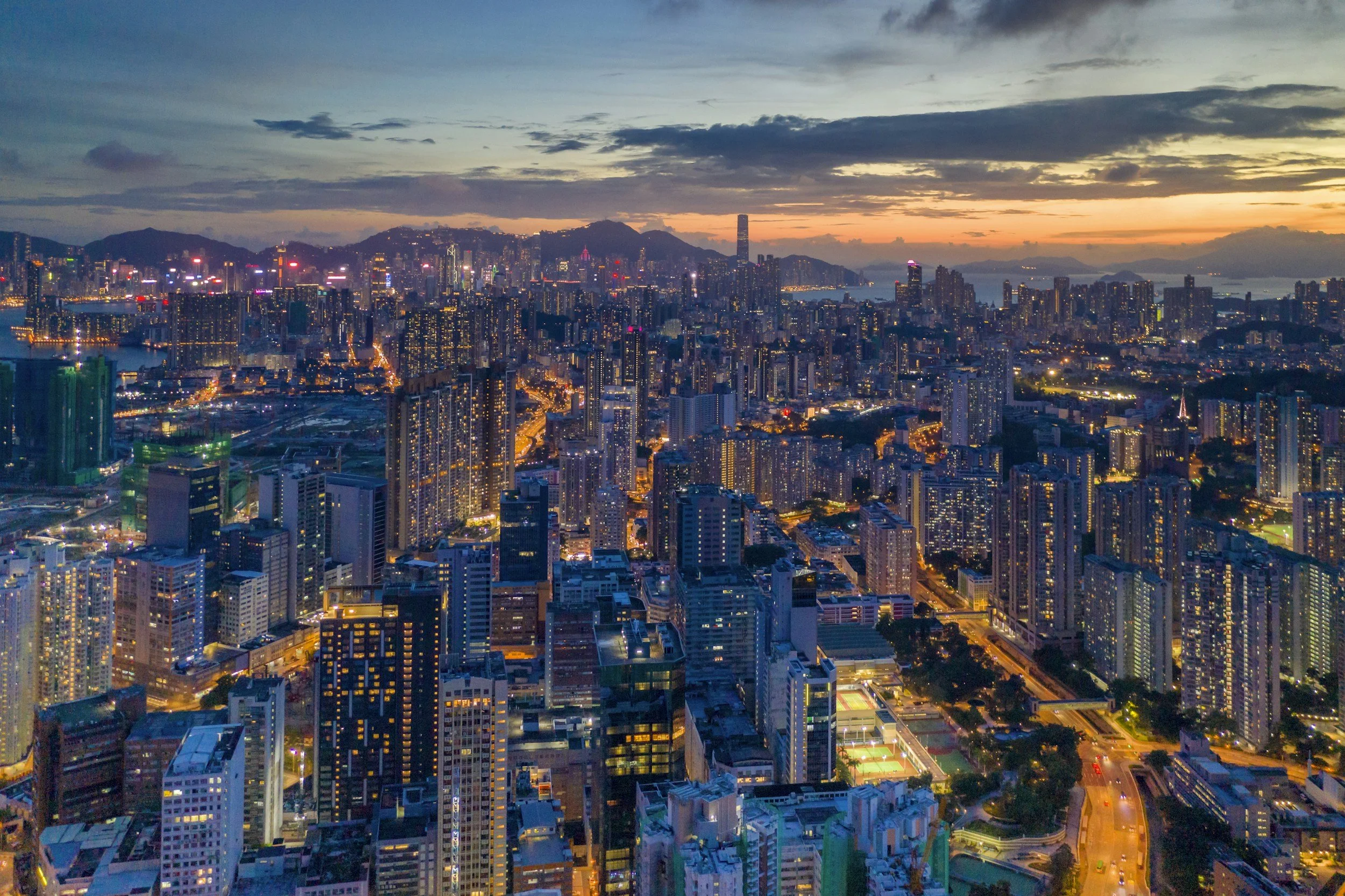 A city skyline at dusk with numerous illuminated tall buildings, mountains in the background, and a partly cloudy sky with orange and blue hues.
