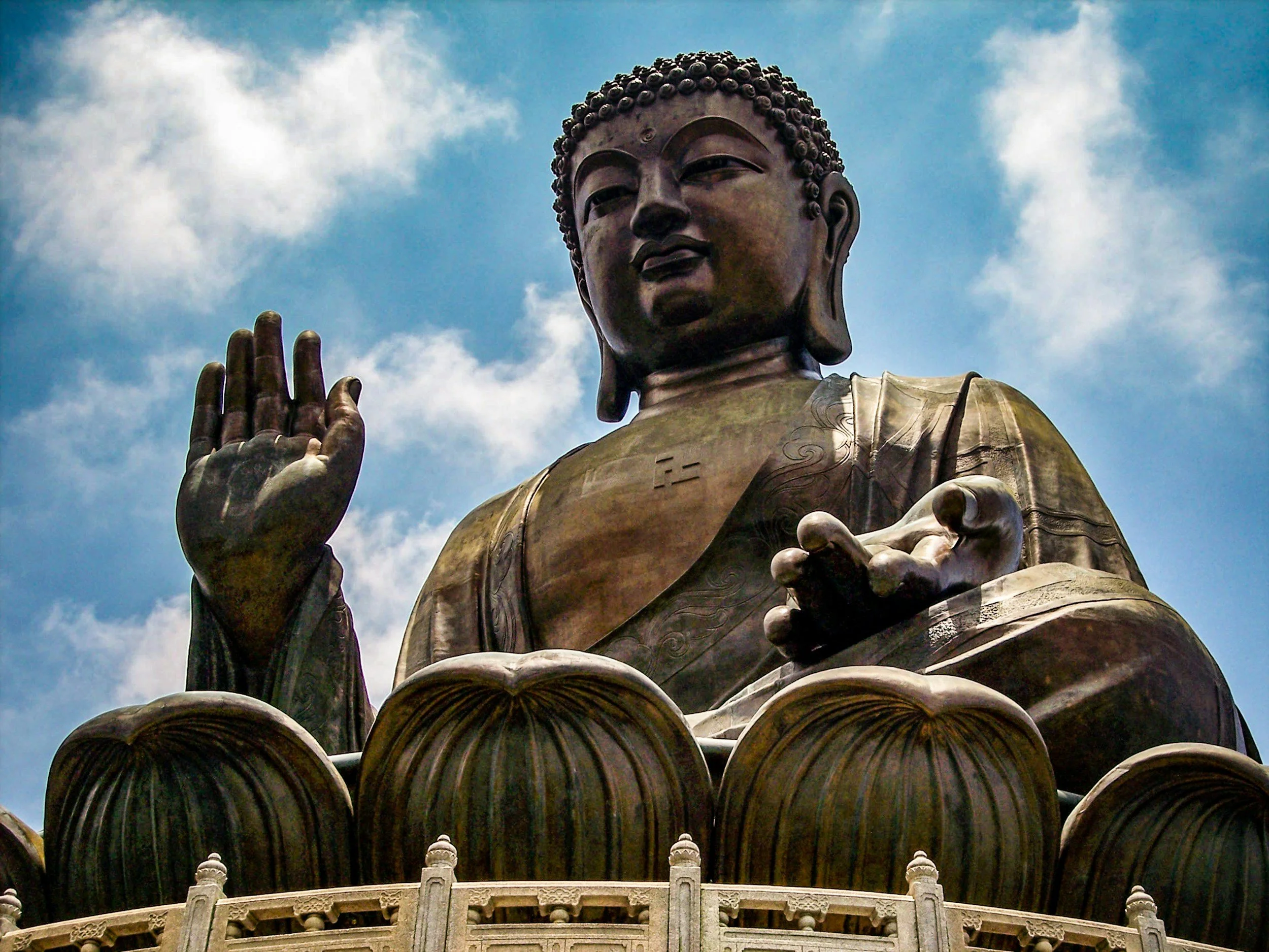 Close-up of a large bronze statue of Buddha with a serene expression and hand raised in a mudra, against a blue sky with clouds.