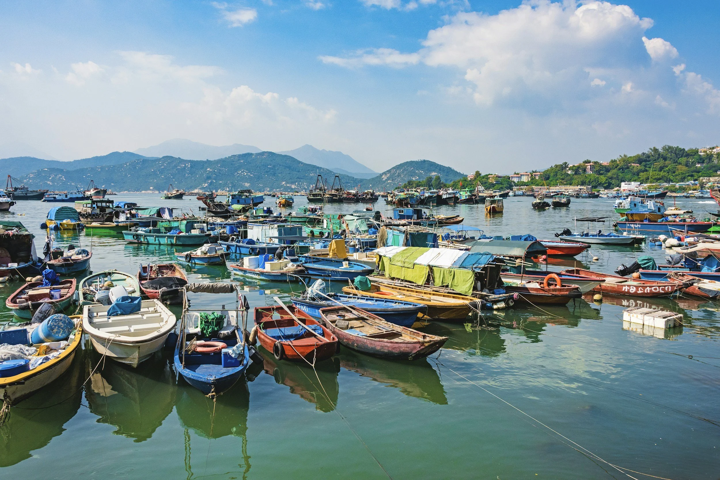 A harbor filled with numerous colorful boats floating on calm water under a partly cloudy sky with mountains in the background.