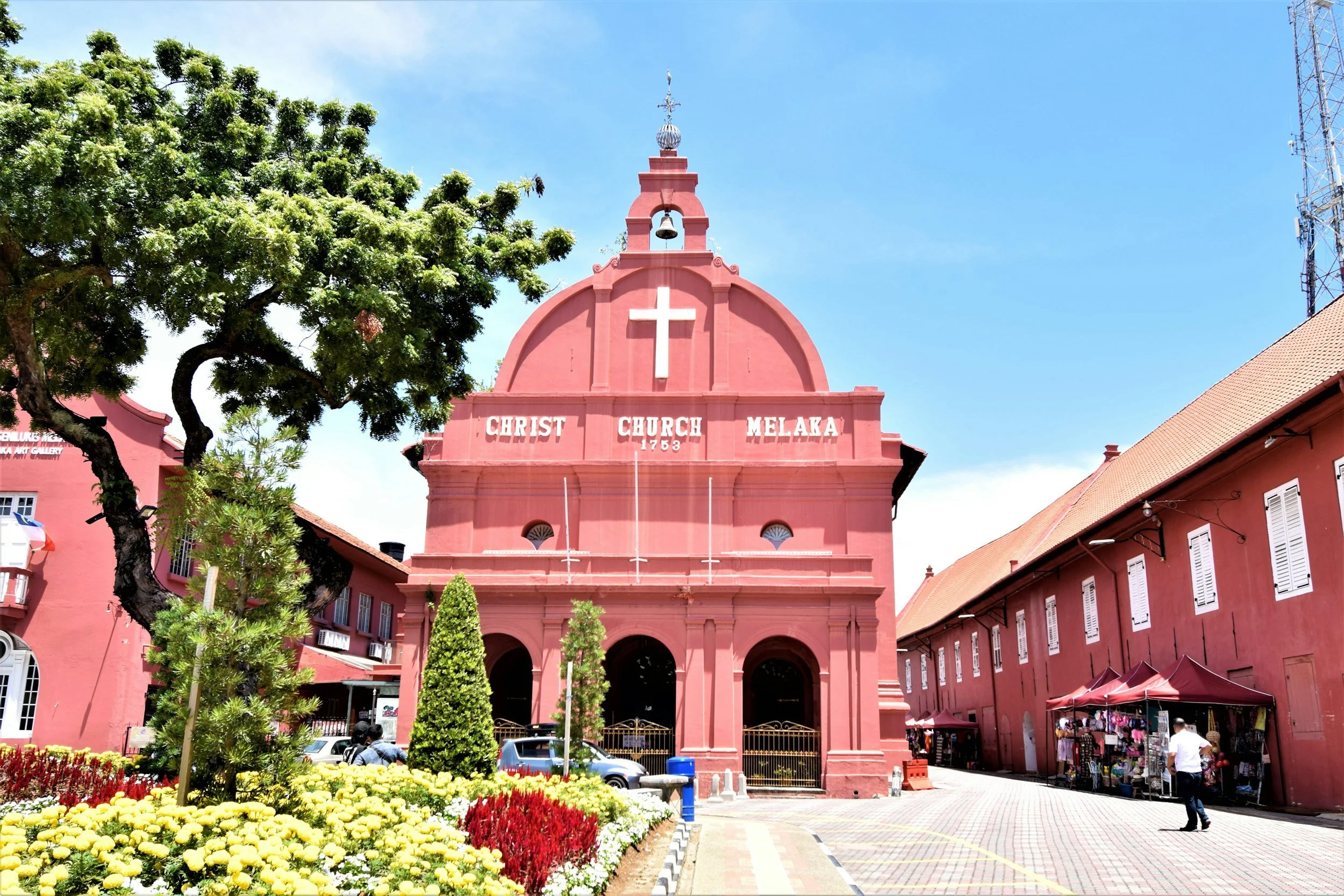 Pink historic church with white cross and bell, FL in Malaysia, surrounded by colorful flowers and trees, with people and market stalls nearby under a clear blue sky.