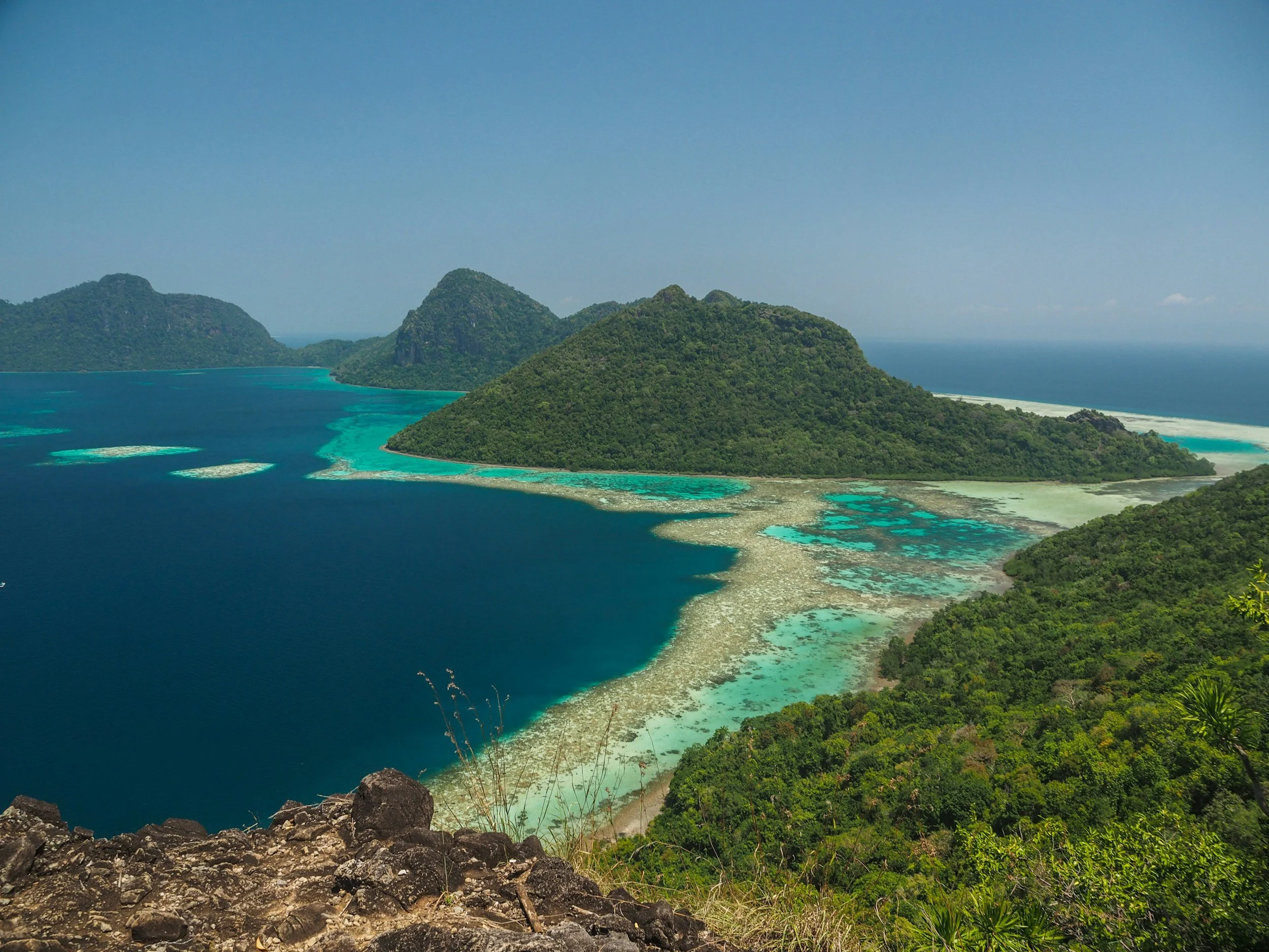A panoramic view of a tropical island with lush green hills, turquoise waters, and coral reefs, seen from a rocky overlook.