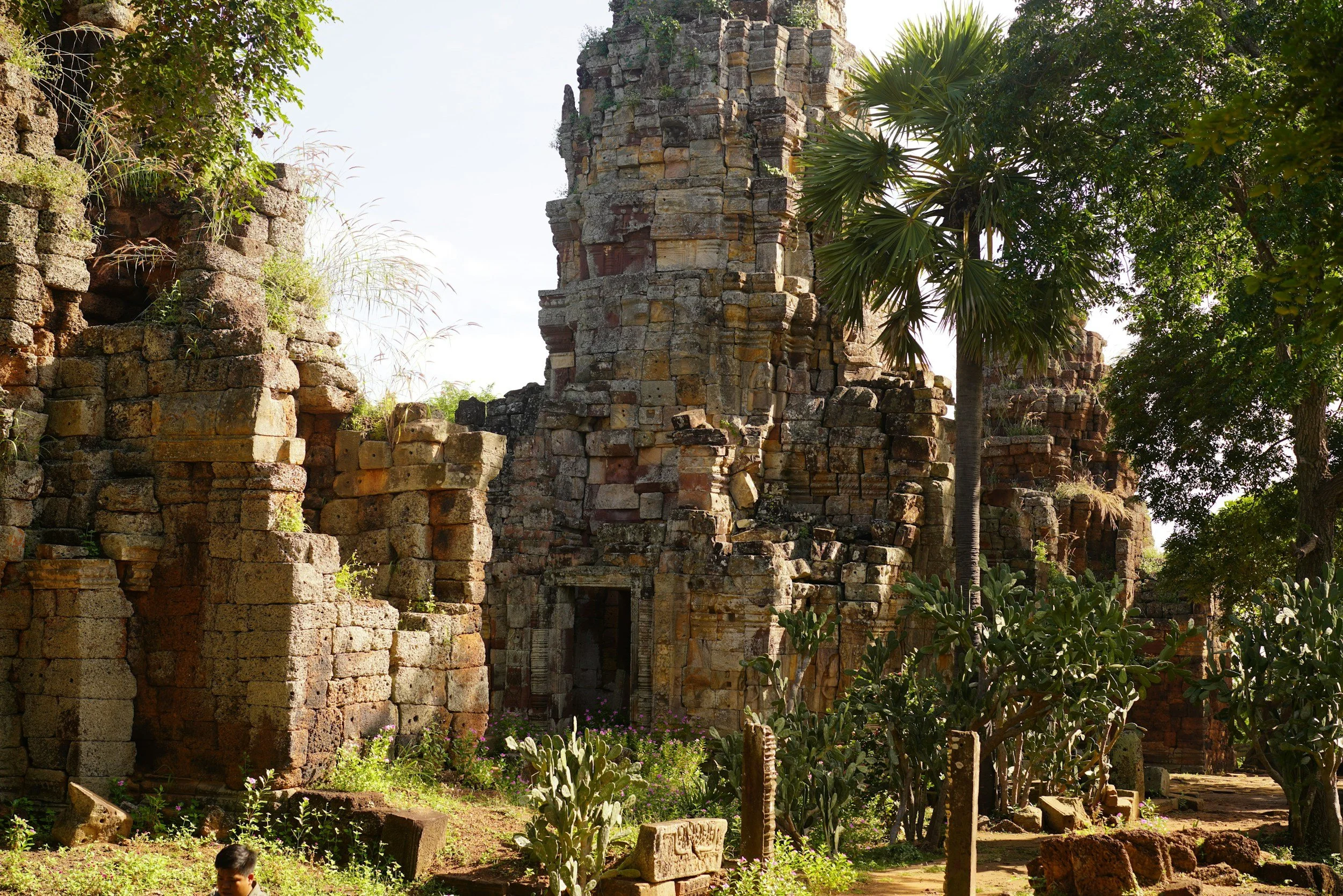 Ancient stone ruins with a tall central structure surrounded by greenery, including trees and cacti, under a bright sky.