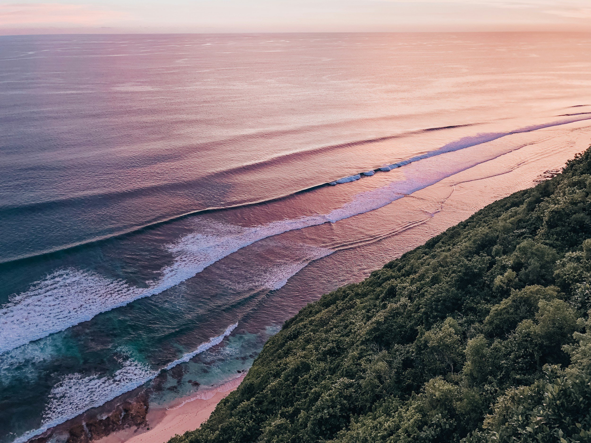 A view of a beach at sunset with gentle waves rolling onto the sandy shore, greenery on a hill in the foreground, and a sky with soft pastel colors.