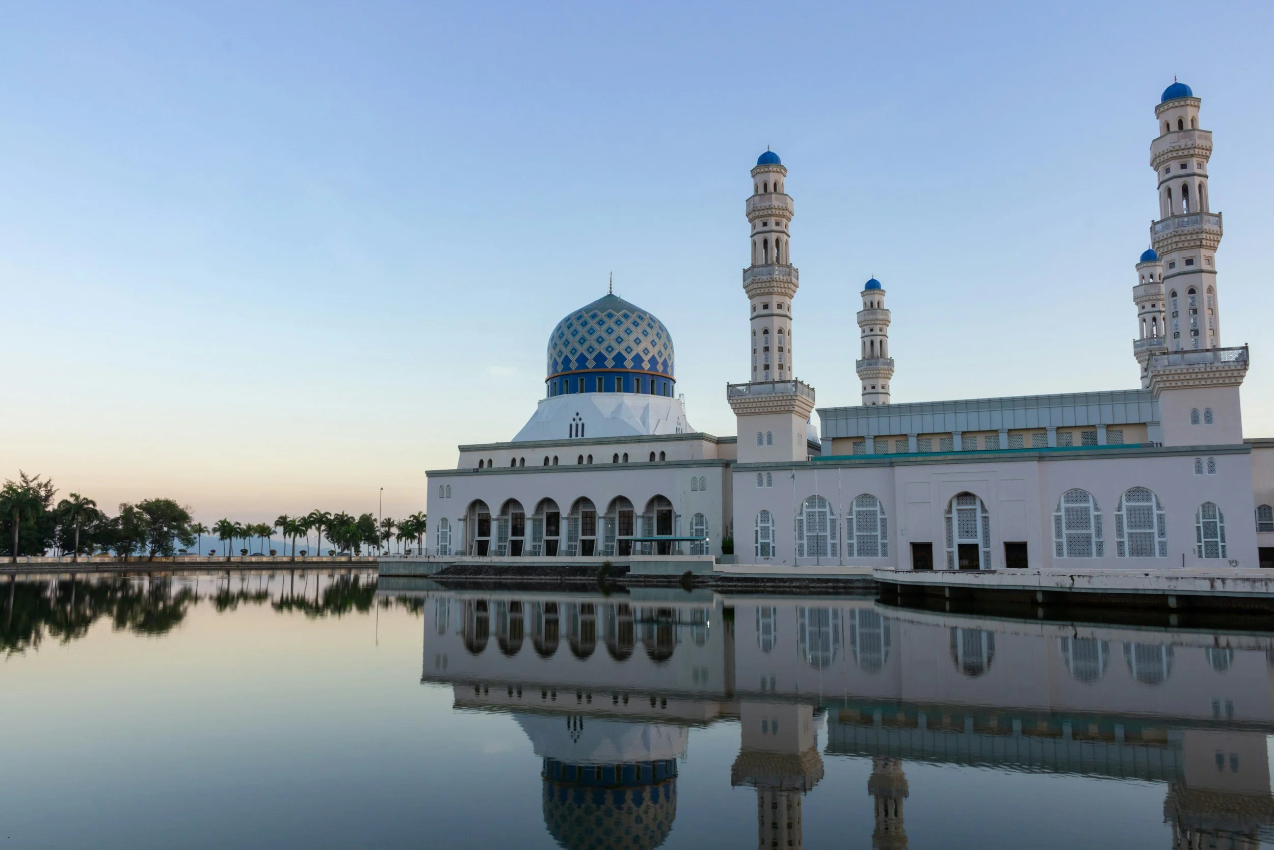 A mosque with a large blue-dome and white minarets reflecting in a body of water, with trees and a clear sky in the background.
