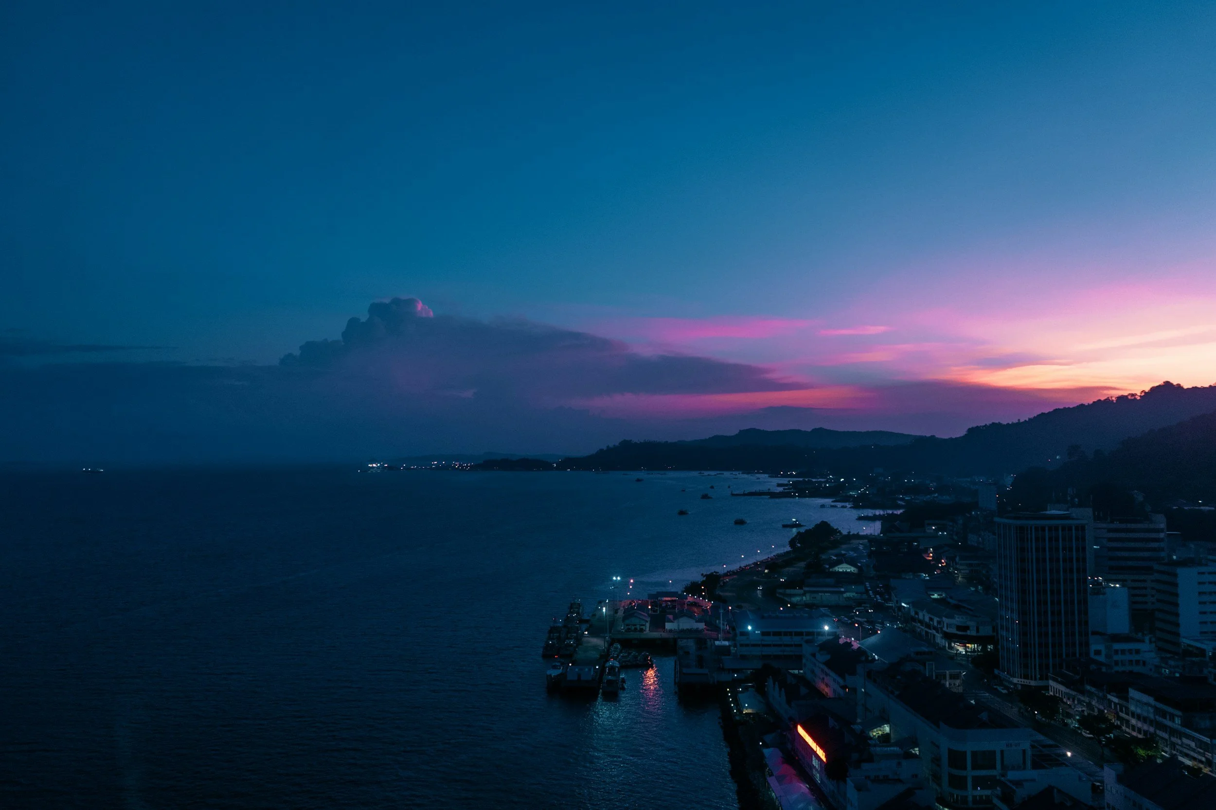 A coastal cityscape during dusk with a vibrant purple and pink sky, city buildings along the shoreline, boats in the water, and mountains in the background.