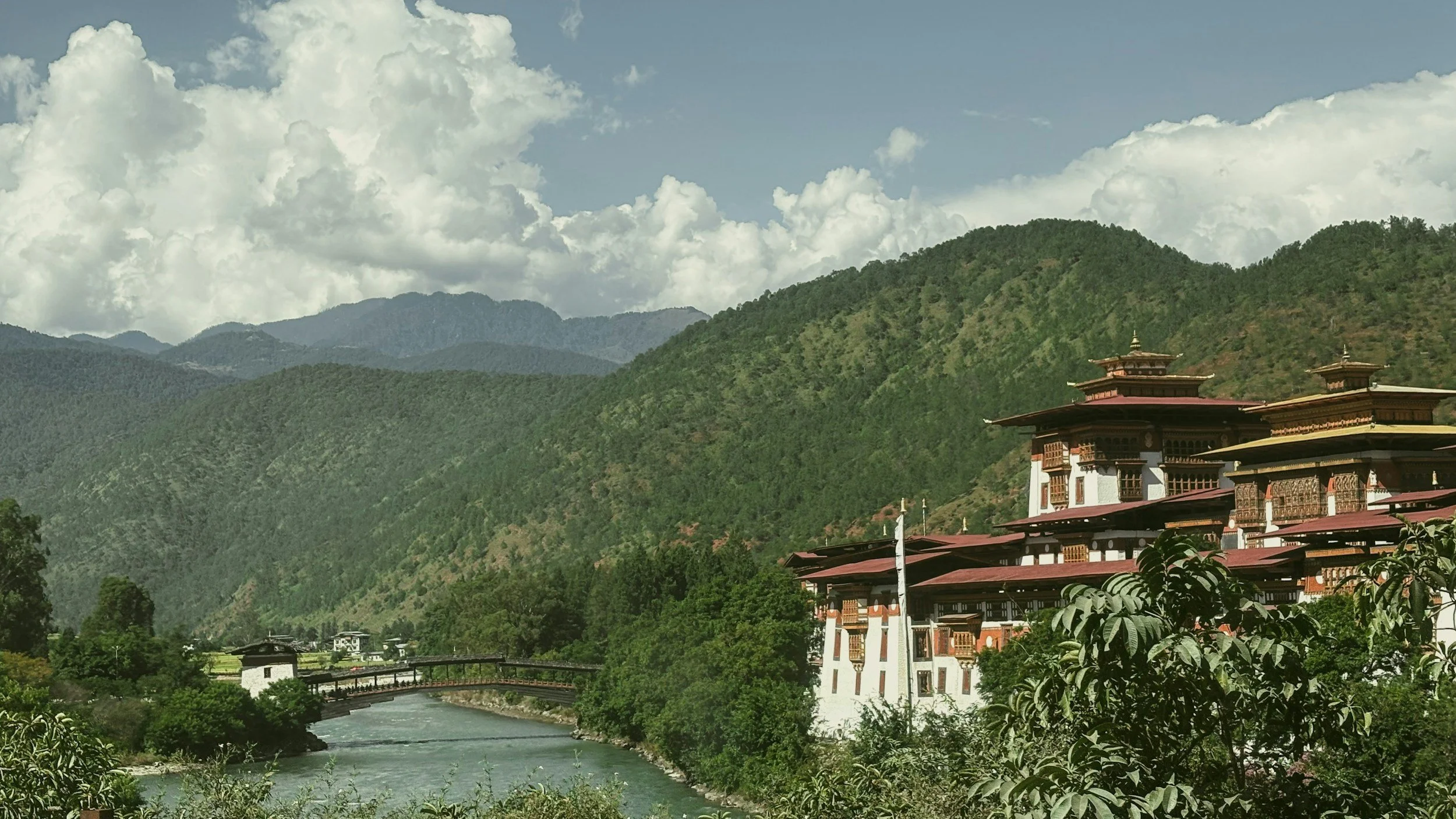 Mountain landscape with lush green hills, traditional building with ornate rooftops, river flowing in the foreground, bridge crossing the river, partly cloudy sky.
