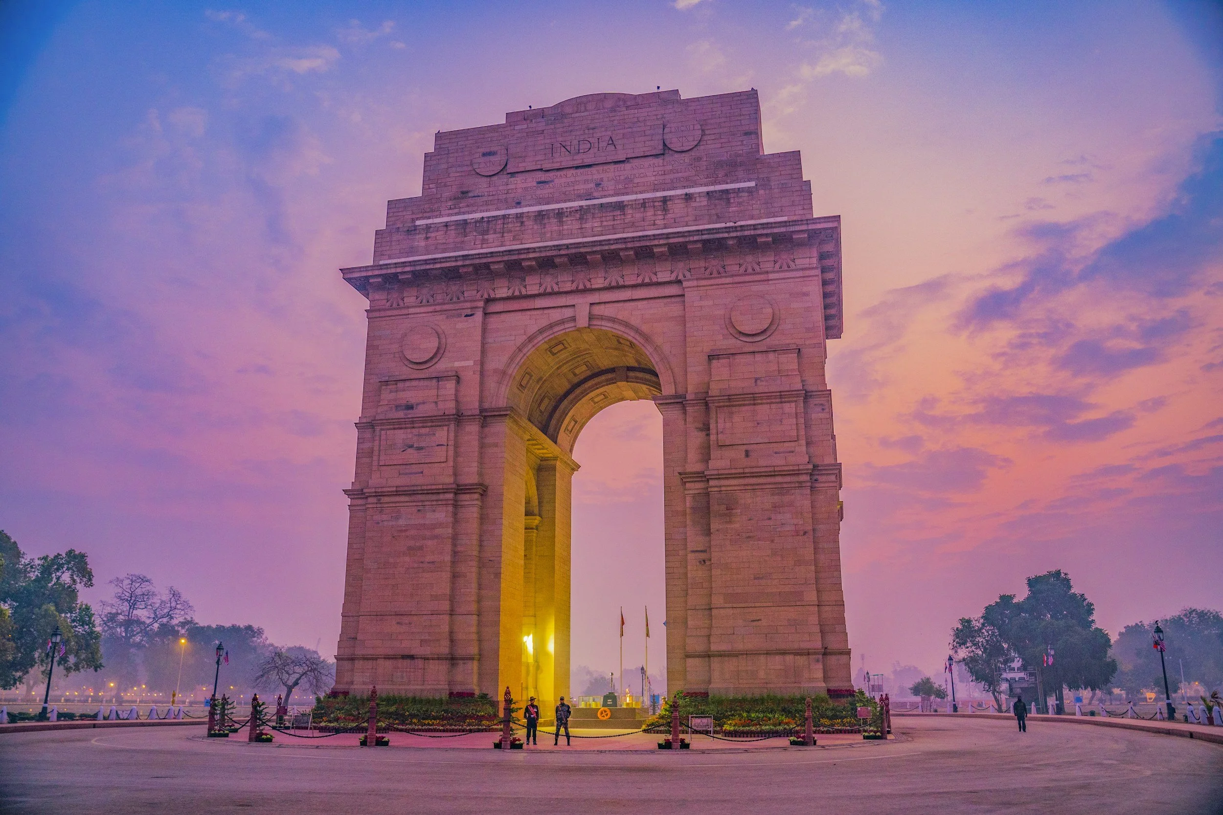 India Gate monument in India illuminated against a cloudy pink and purple sky during sunset, with people walking nearby.