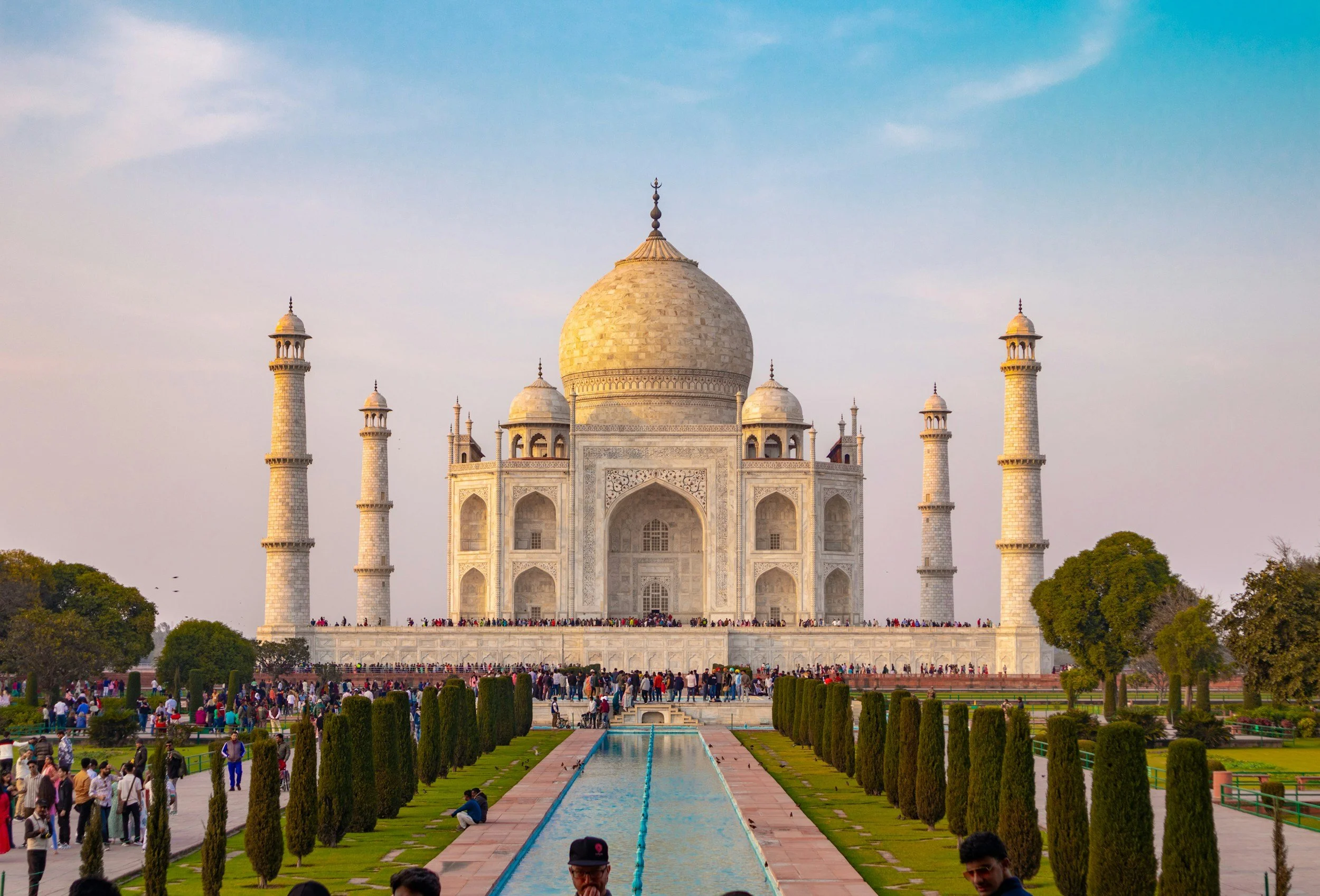 Photo of the Taj Mahal in India, with tourists in the foreground and lush gardens. The monument is a white marble mausoleum with a large central dome and four minarets.