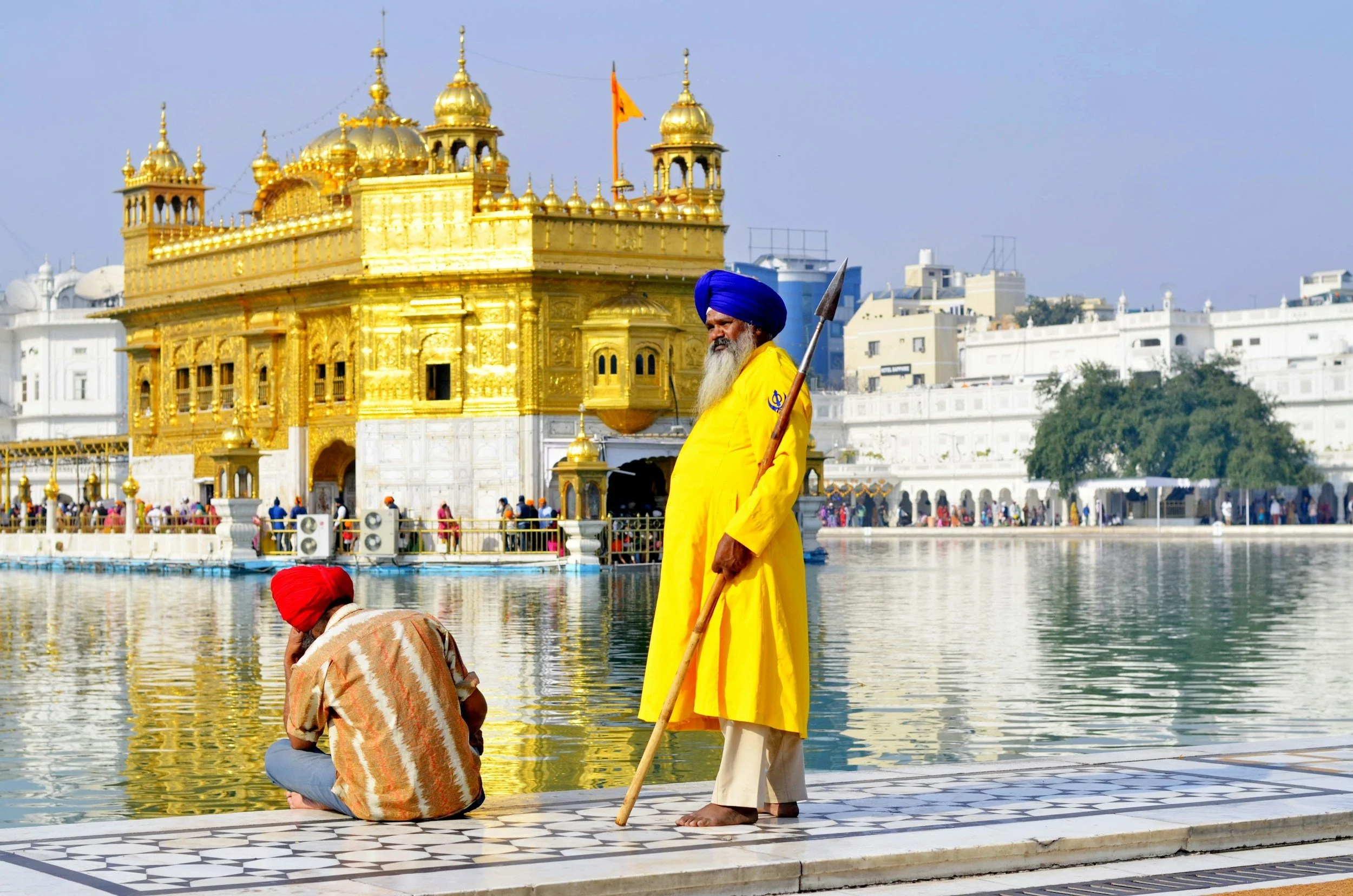 A golden temple with intricate architectural details located near a body of water, with a man wearing a yellow garment and blue turban standing and holding a spear, and another person sitting by the water wearing a red turban