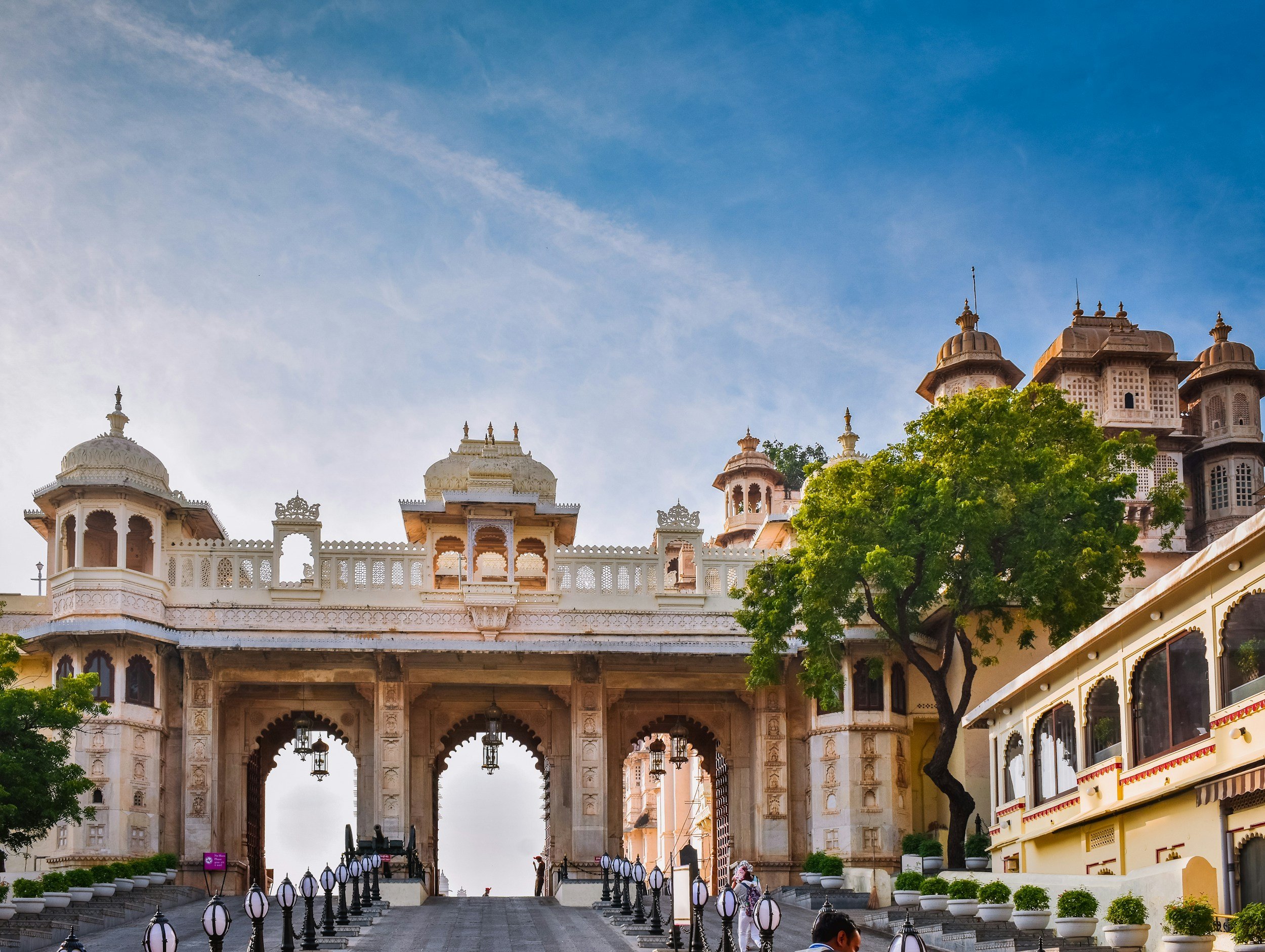 A grand white architectural building with Indian design elements, featuring multiple domes, arches, and intricate details, set against a clear blue sky with some green trees nearby.