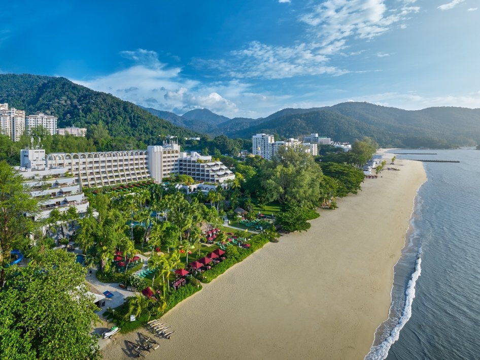 A scenic beach resort with white sandy beach, green trees, lush landscaping, and multiple resort buildings against a backdrop of mountains and blue sky.