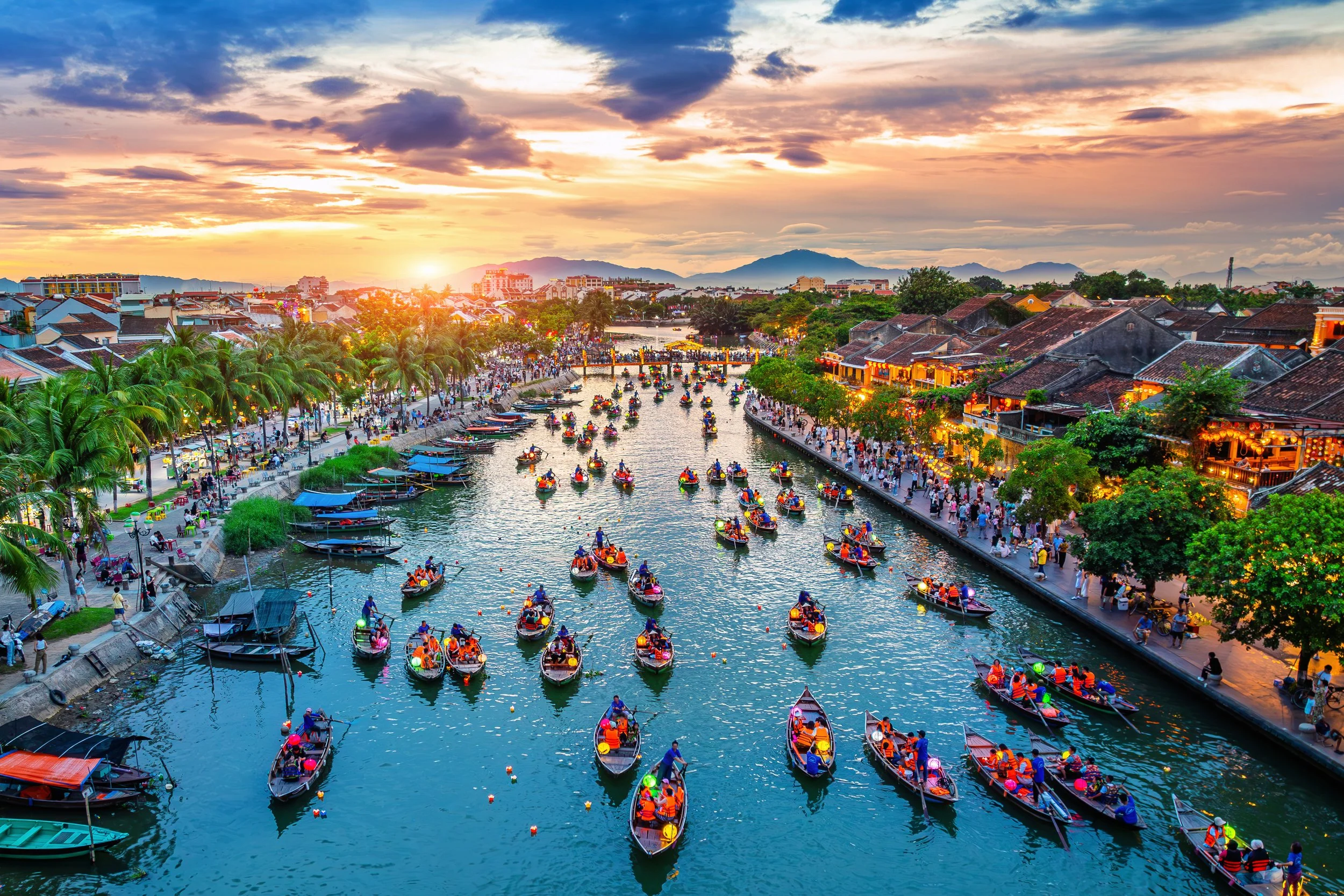 Sunset over a lively river scene with numerous small boats filled with people, lined by a busy promenade with greenery and buildings, amidst mountains in the distance.