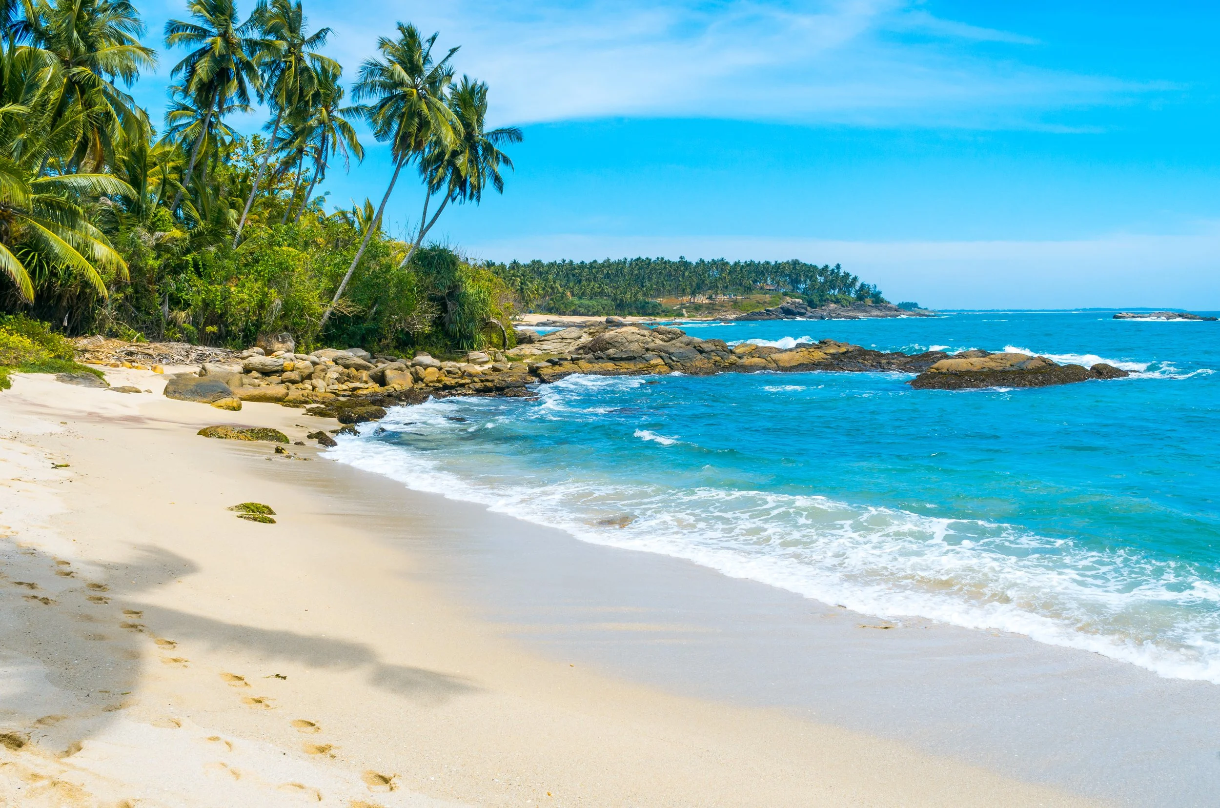 Tropical beach with white sand, rocky shoreline, lush green palm trees, and bright blue ocean with waves under a clear blue sky.