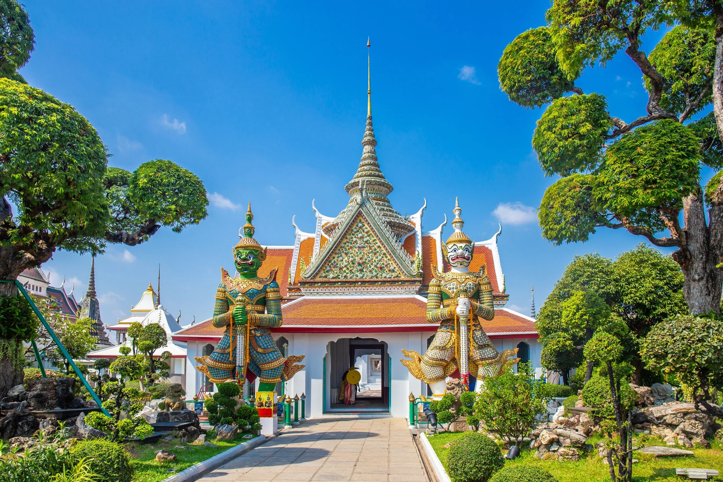 A traditional Thai temple with ornate architecture, two giant guardian statue figures flanking the entrance, and lush green trees surrounding the area under a bright blue sky.