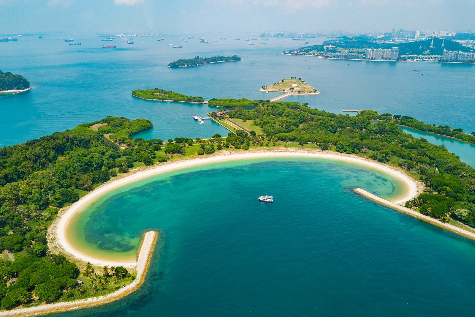 Aerial view of a lush green island with a curved sandy beach and calm turquoise waters, surrounded by boats and water.