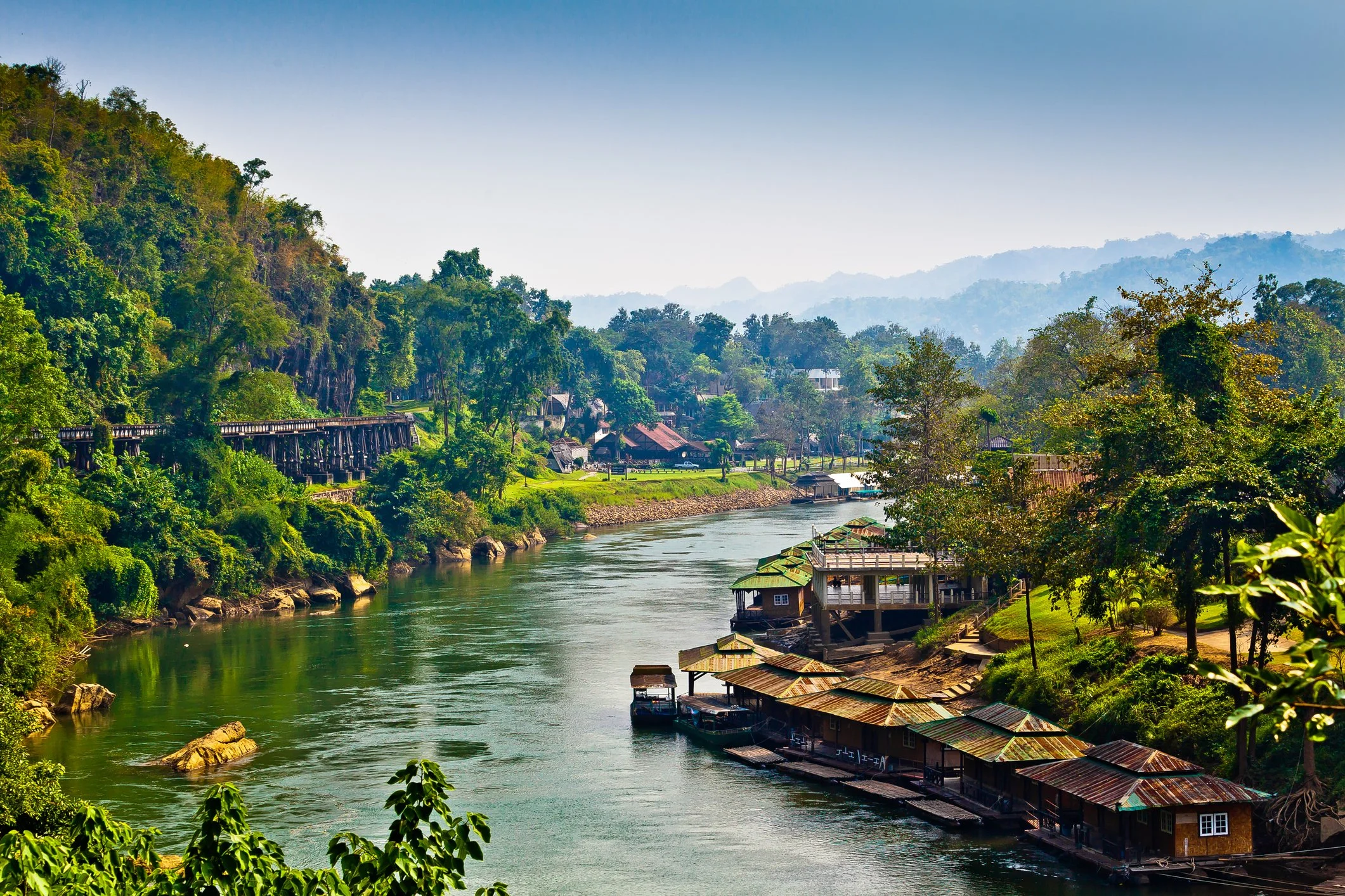 A riverside village with houses on stilts, surrounded by dense green forest, with a bridge crossing the river and mountain hills in the background.