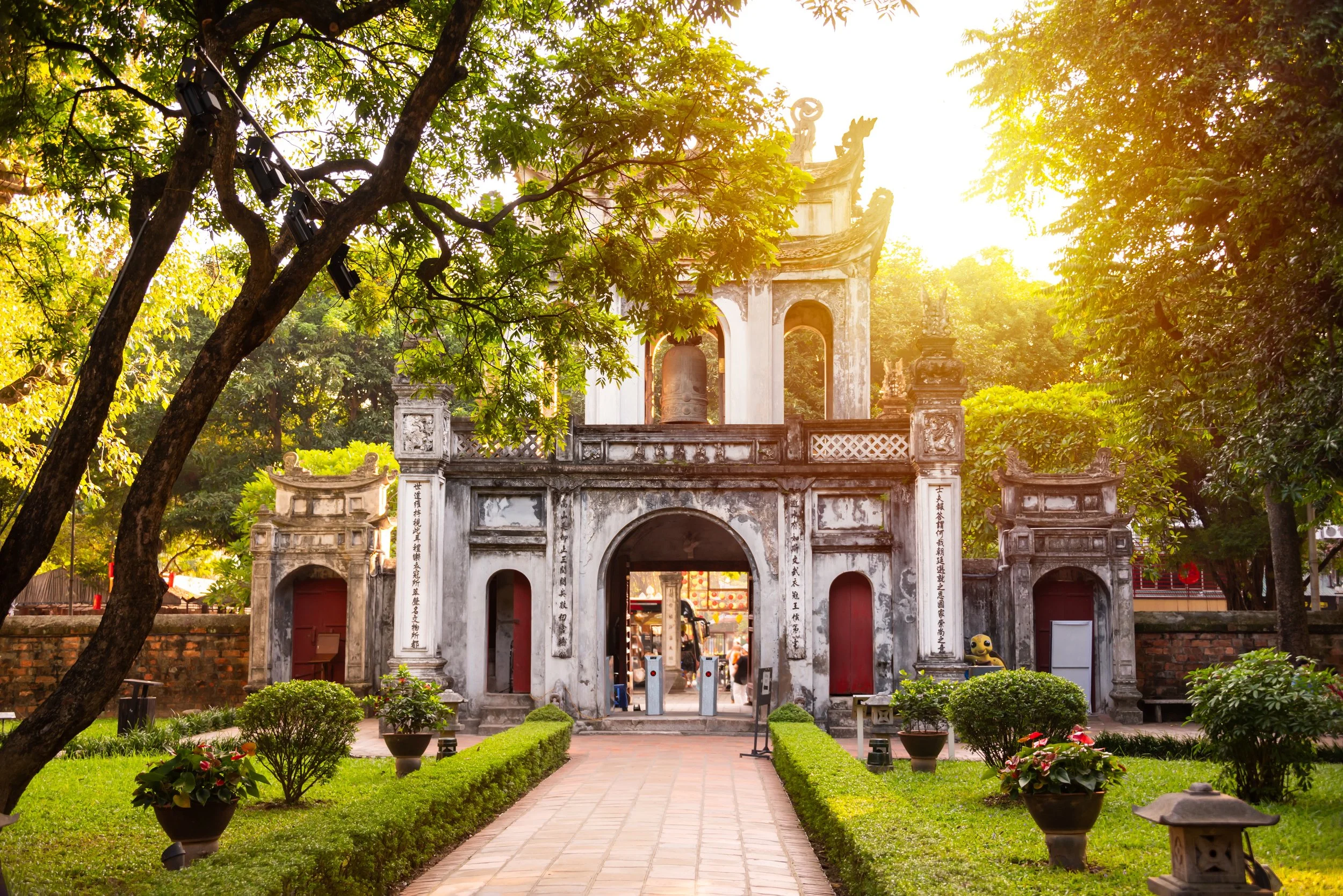 An entrance to a historical or cultural site with an archway, surrounded by trees and a garden, illuminated by warm sunlight.