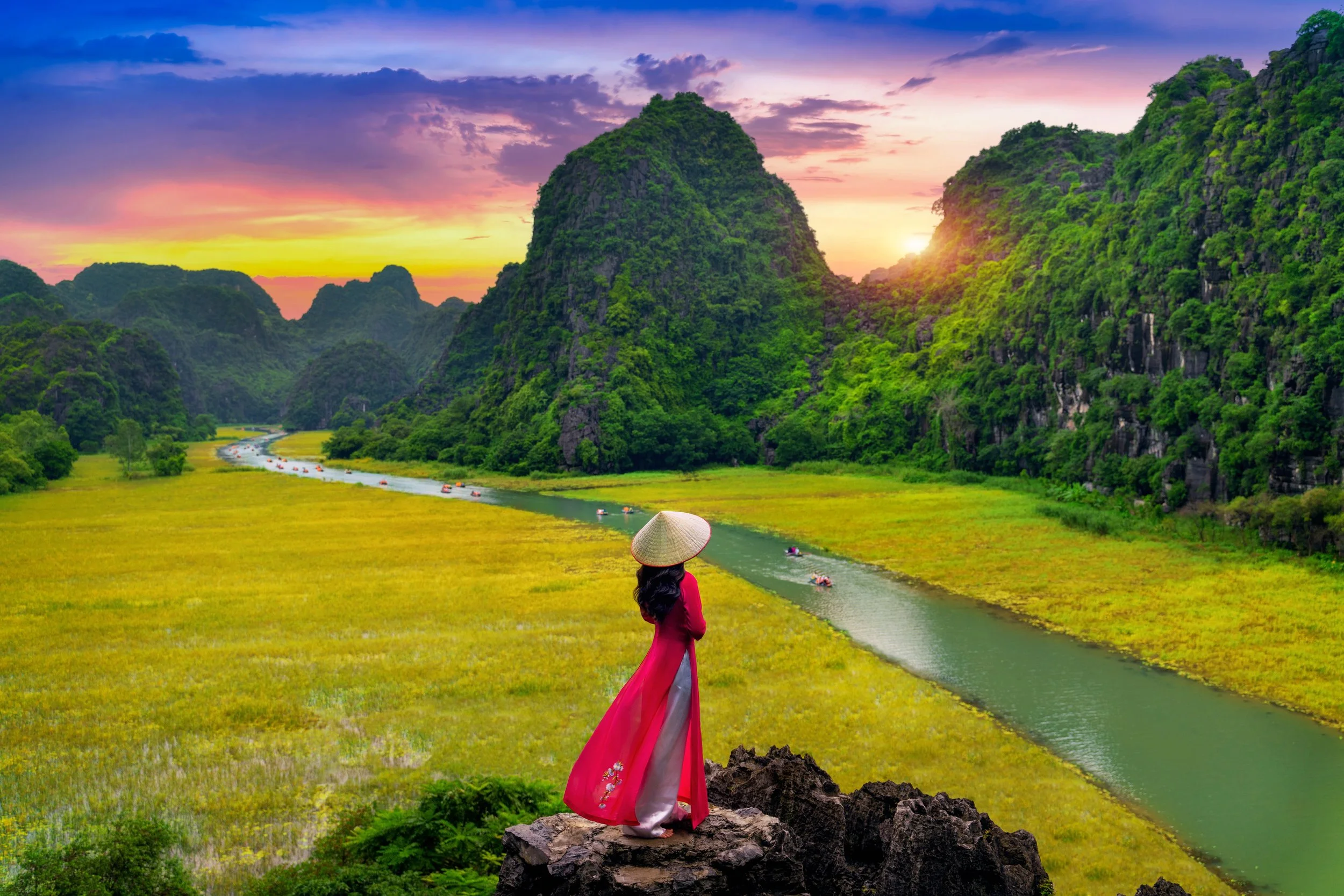 A woman dressed in traditional Vietnamese clothing, standing on a rock, overlooking a river surrounded by lush green mountains at sunset.