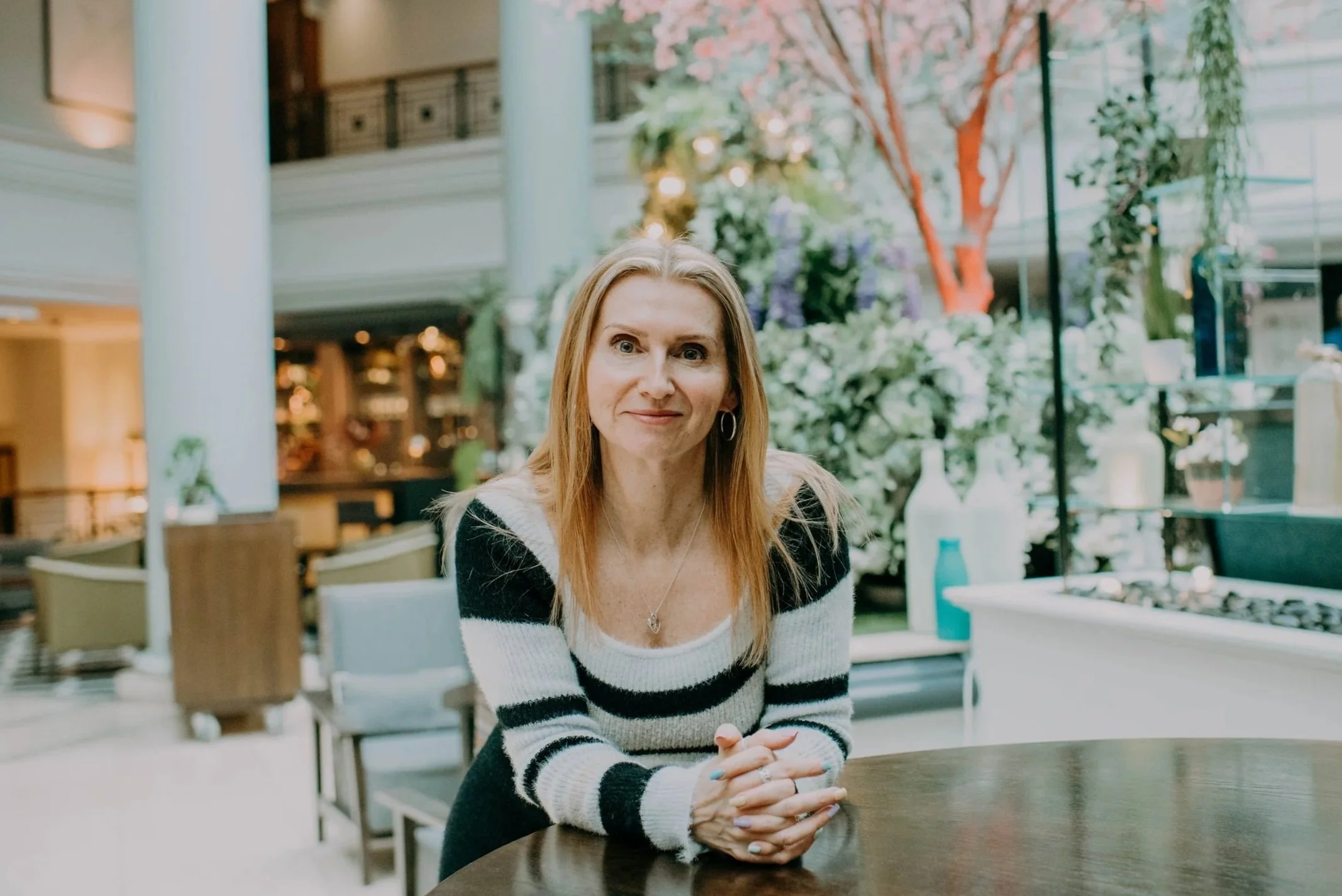 A woman with red hair and blue eyes sitting at a round table in a bright, modern indoor space with plants and trees in the background.