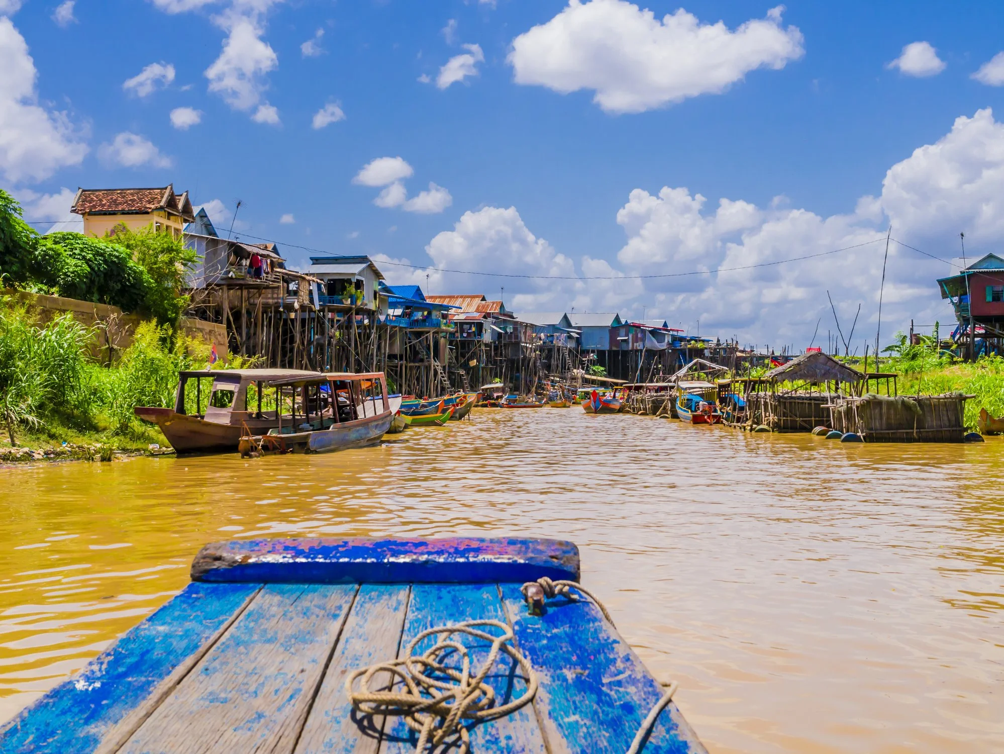 View from a boat on a river with houses on stilts along the shoreline, boats docked by the water, and a bright blue sky with scattered clouds.