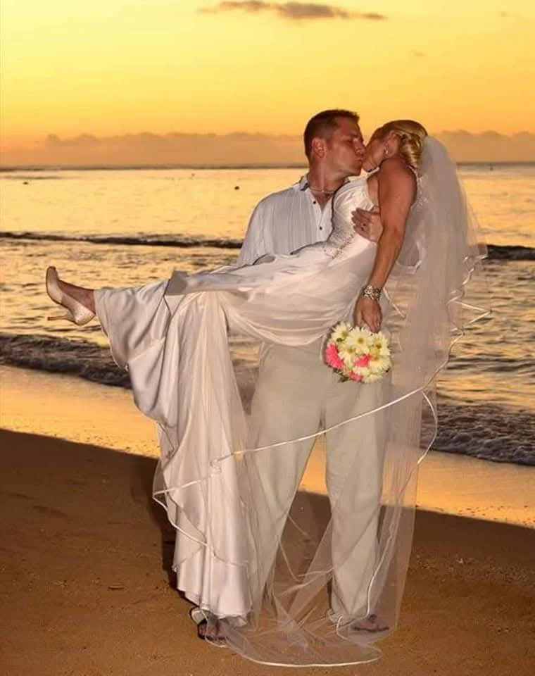 A wedding couple sharing a kiss on the beach during sunset. The groom is holding the bride in his arms, and the bride holds a bouquet of flowers.