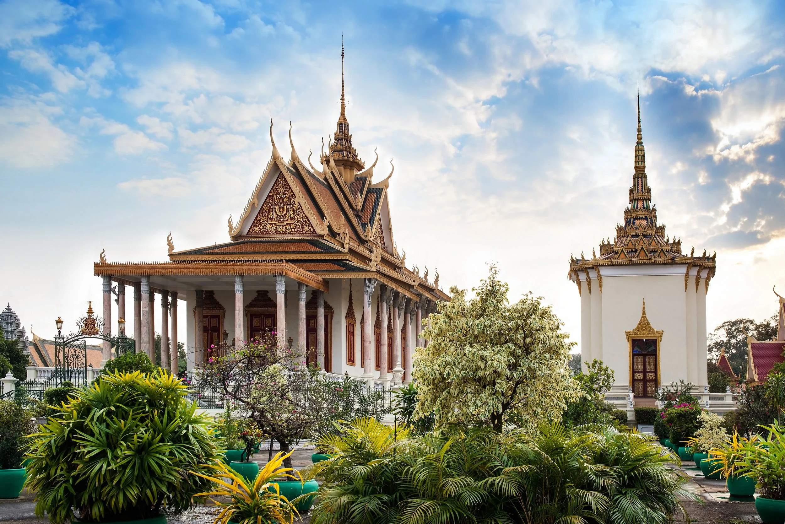 A traditional ornate Thai temple with intricate roof designs and spires, surrounded by lush green plants and trees, under a bright sky with scattered clouds.