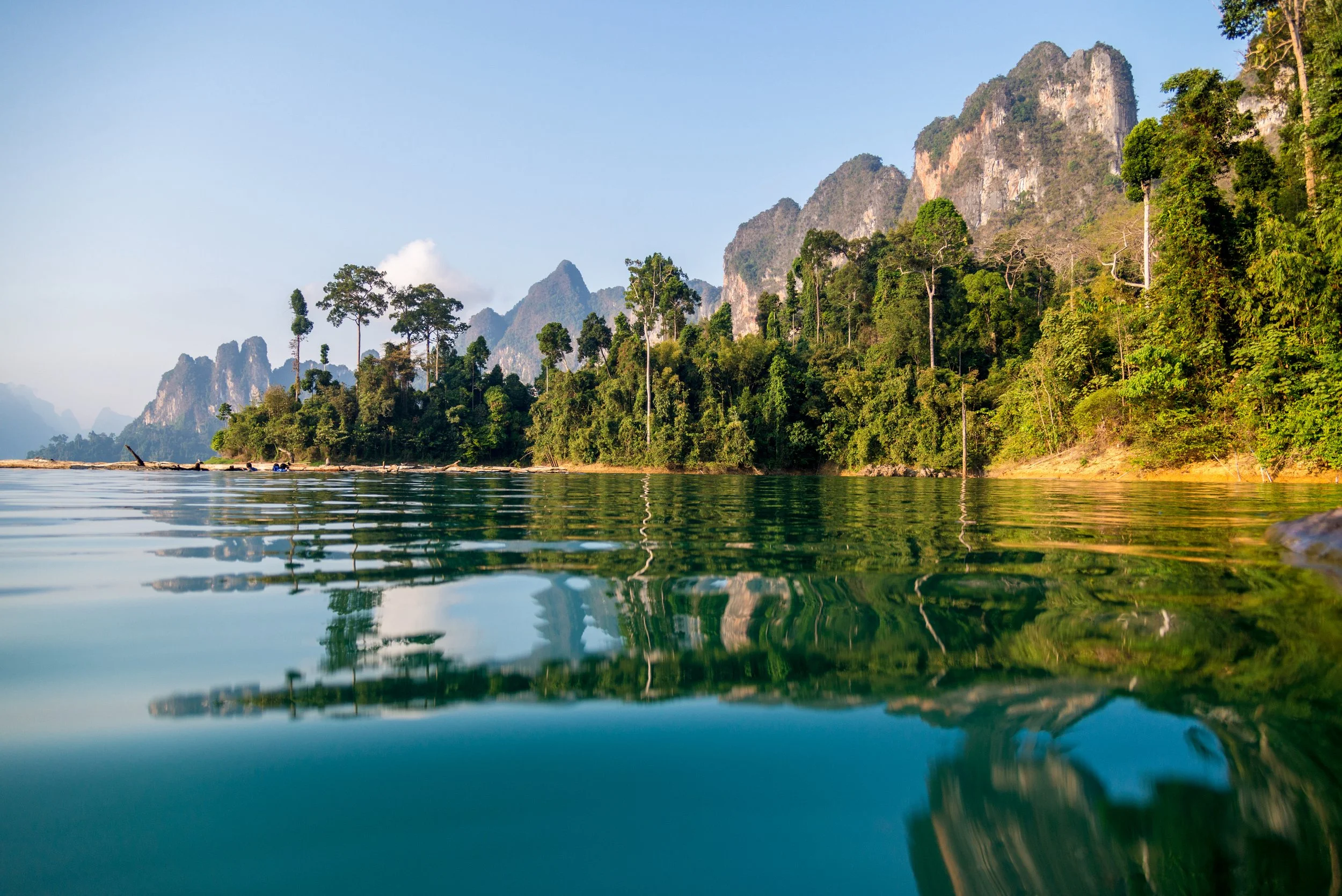 A scenic view of a river with calm water reflecting lush green trees and towering mountains in the background, under a clear blue sky.