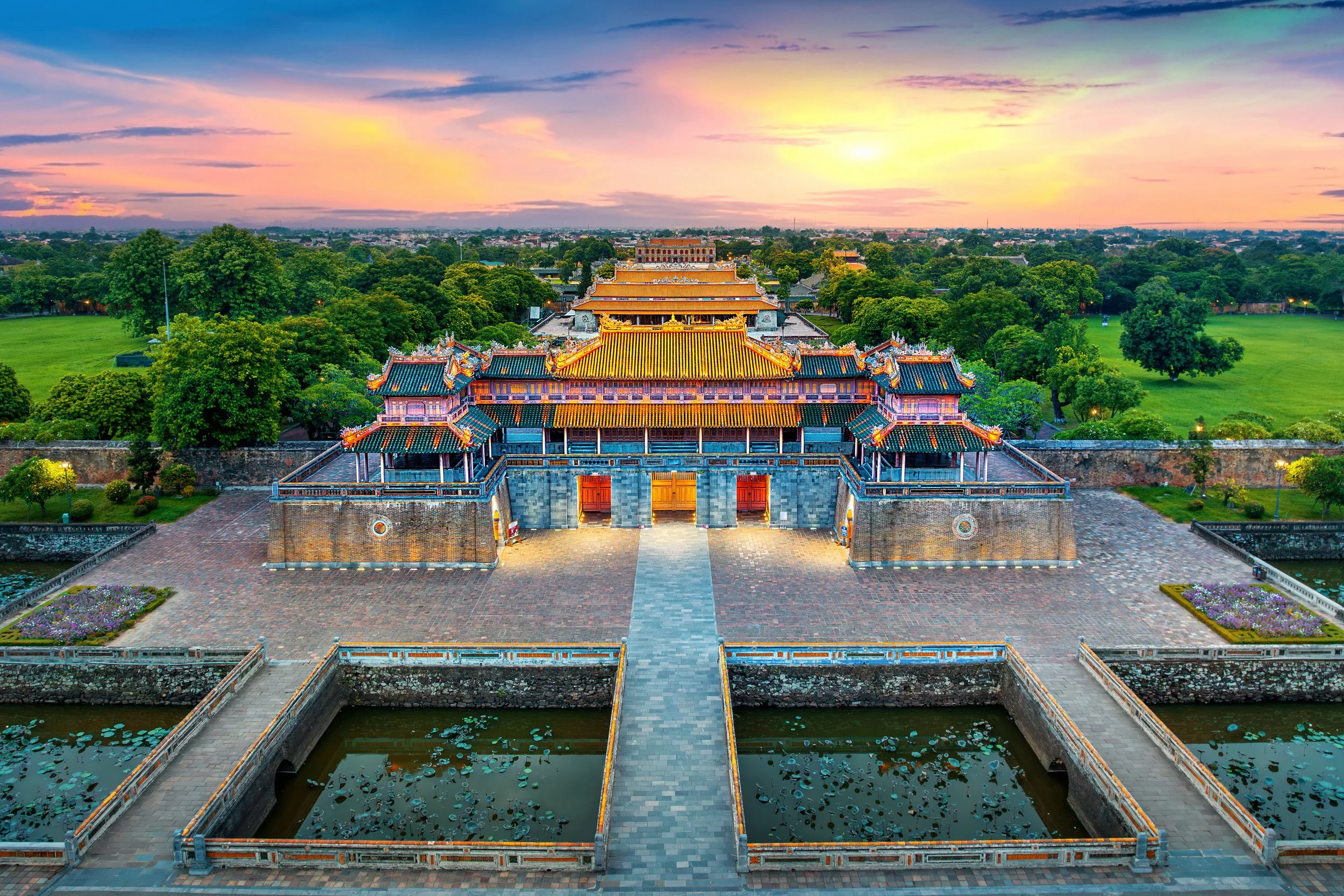 A colorful temple complex with traditional Asian architecture, surrounded by lush green trees and a body of water in the foreground, under a vibrant sunset sky.