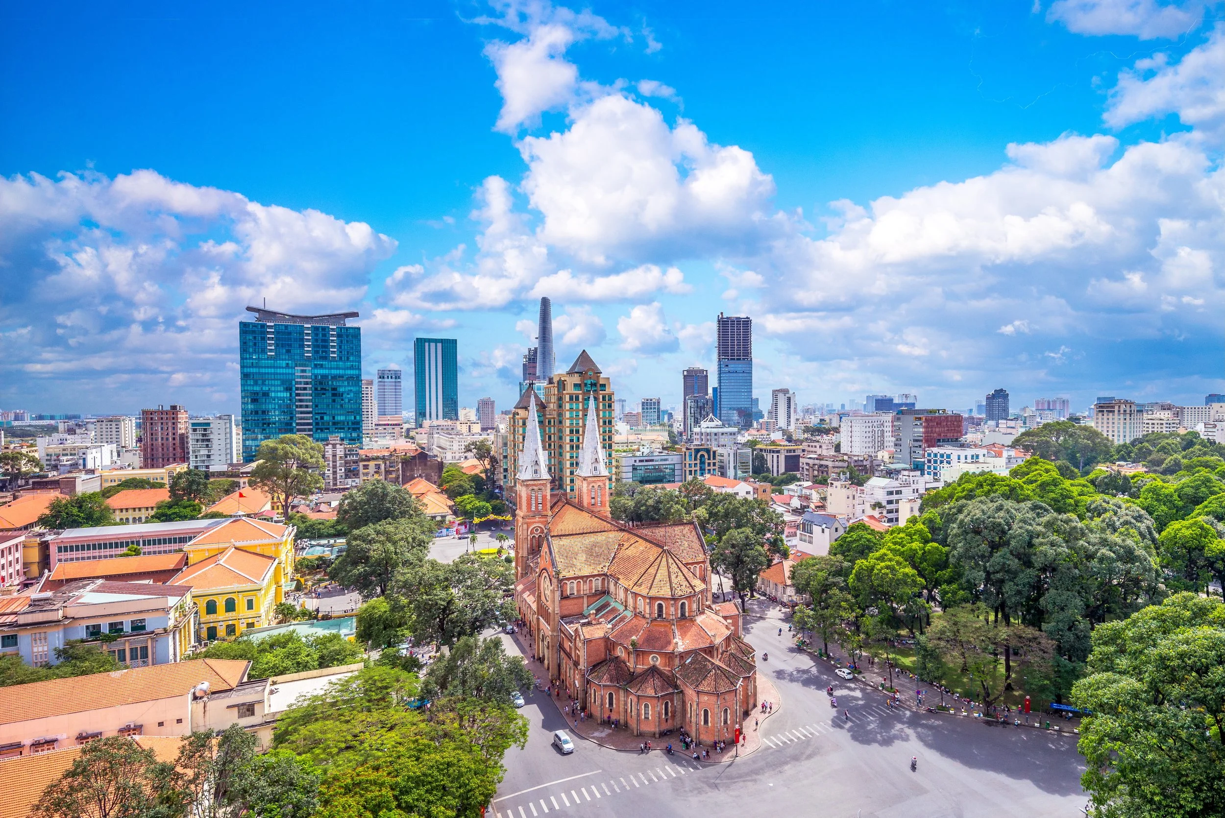 A city skyline featuring modern skyscrapers in the background and a historic church with reddish-brown brickwork and two white steeples in the foreground, surrounded by trees and streets.