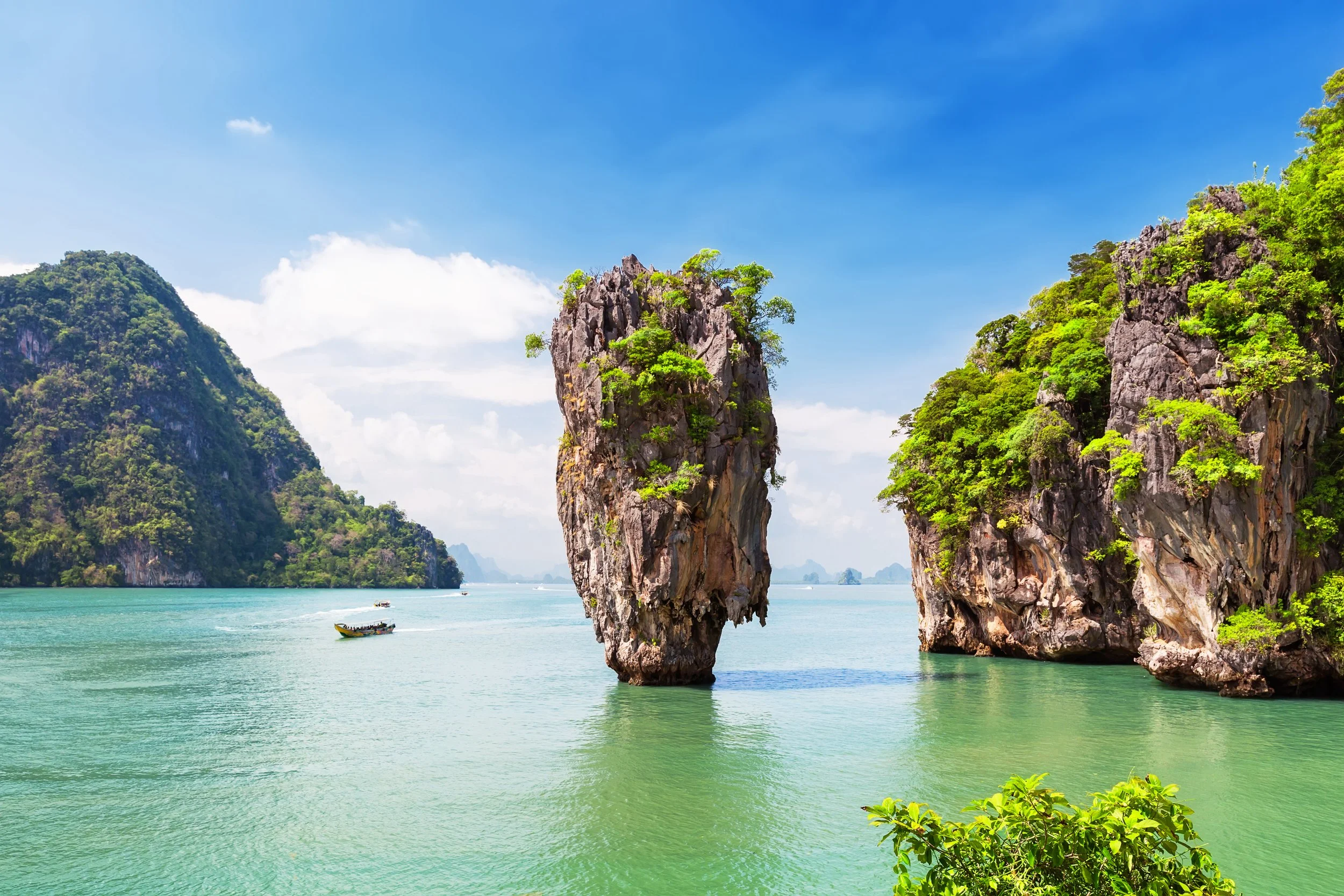 Limestone isles with green foliage rising from turquoise waters under a partly cloudy sky, with boats in the water.
