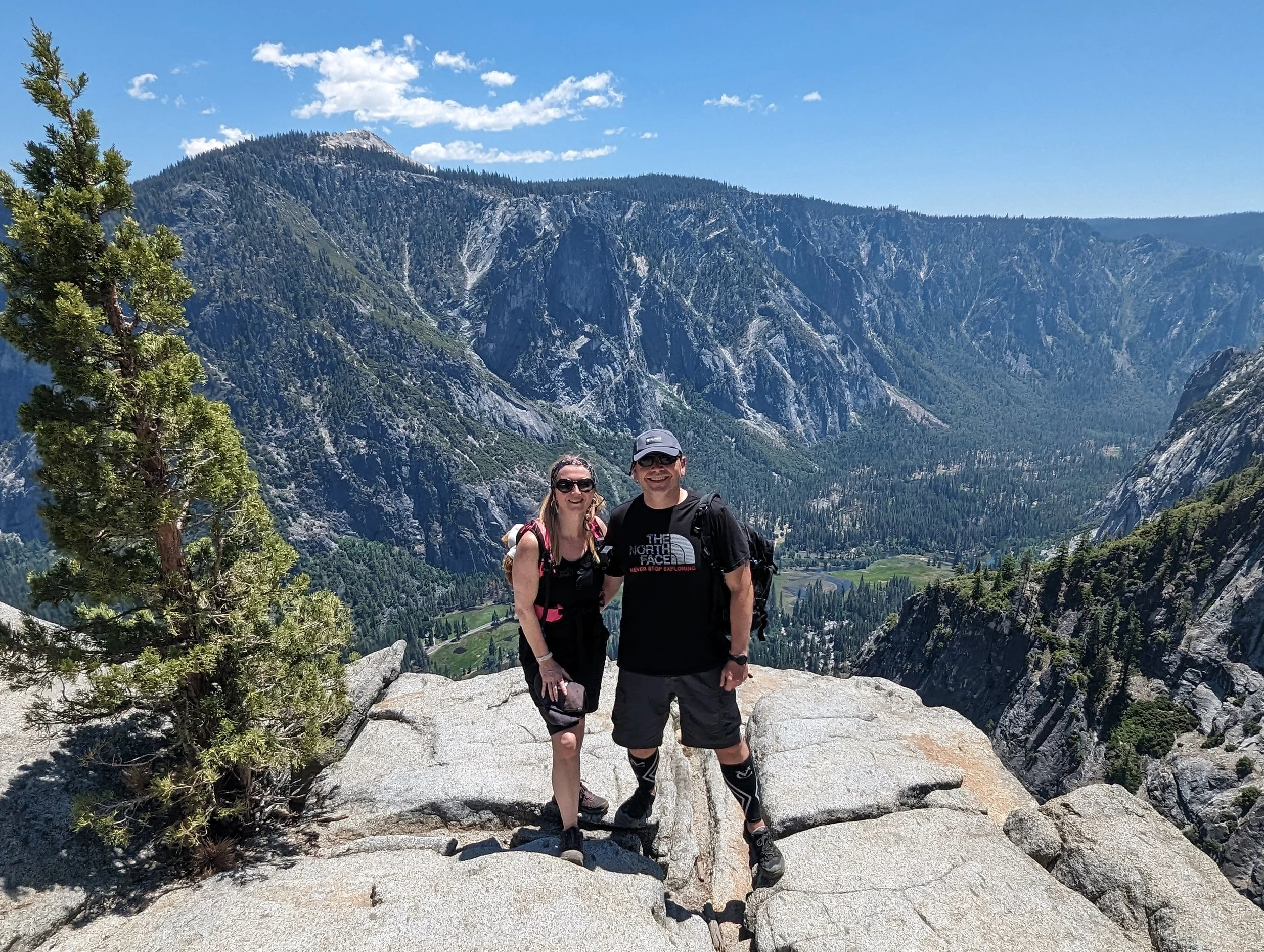 A smiling couple standing on a rocky ledge in a mountainous area with a deep valley, tall pine trees, and rugged cliffs in the background under a clear blue sky.