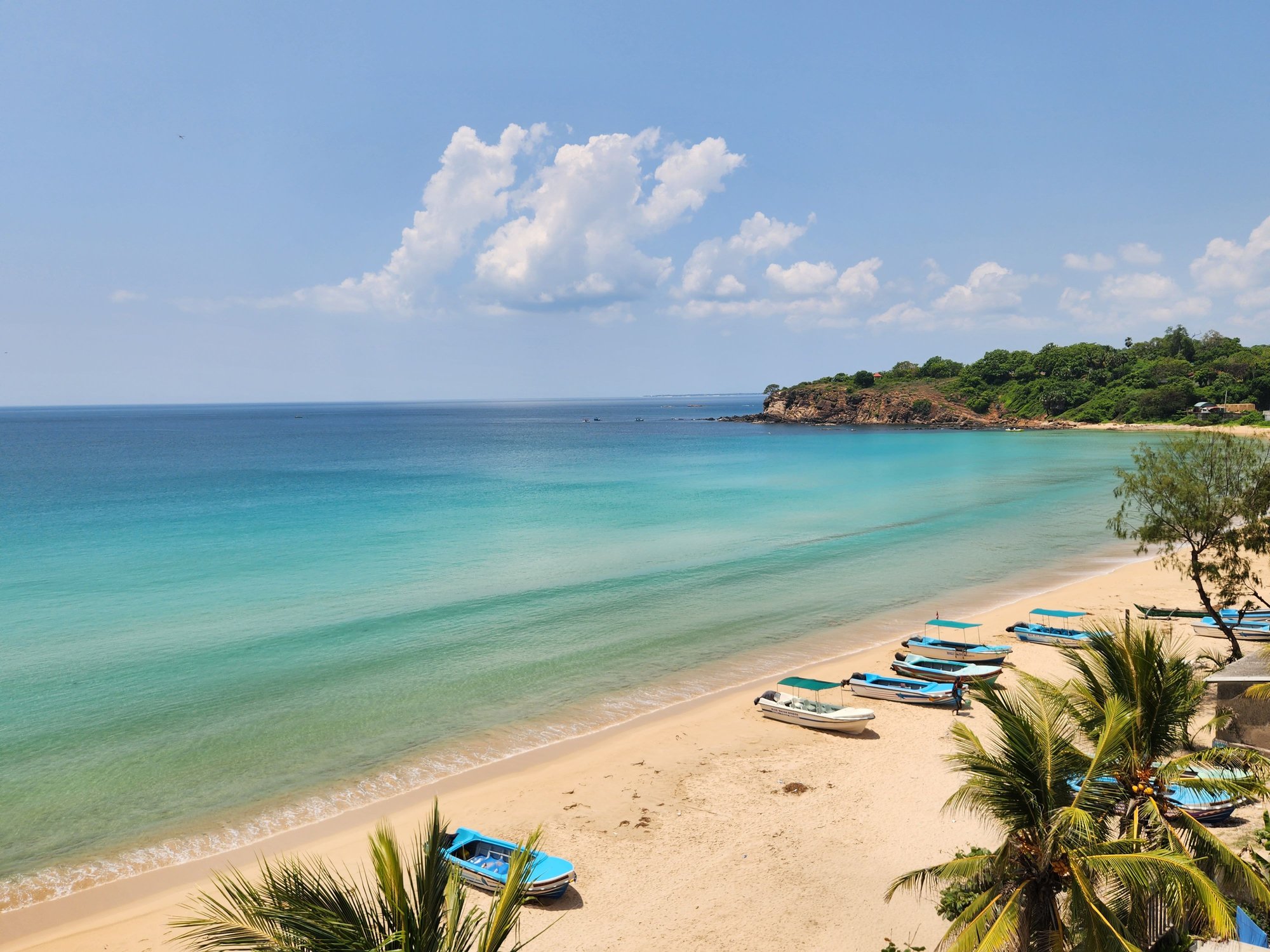 A scenic beach with turquoise water, soft sandy shore, colorful boats, lush green trees, and a hillside with houses under a partly cloudy sky.