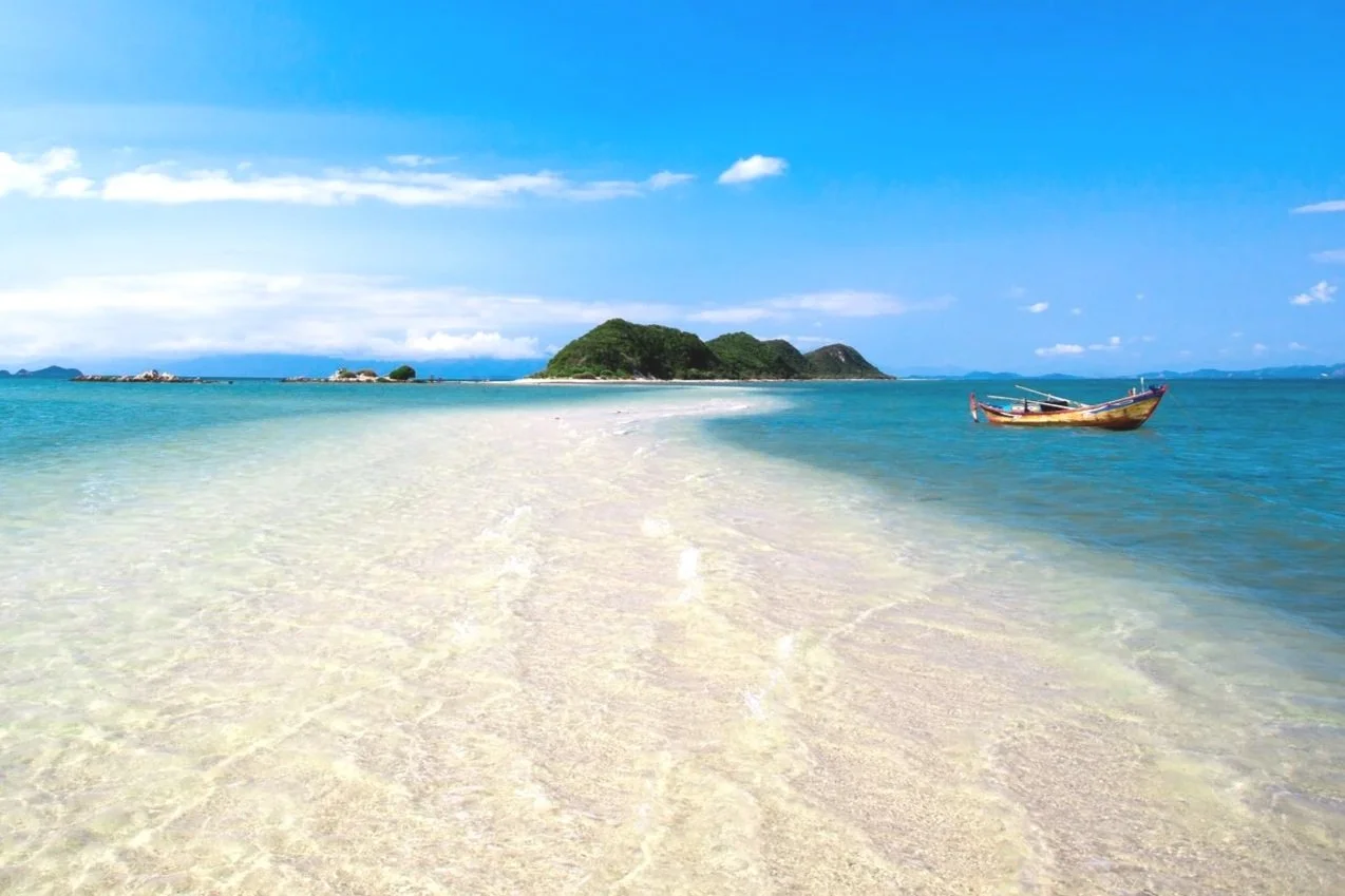 A tropical beach with white sand and clear blue water, a small boat floating near the shore, and green islands in the distance under a partly cloudy sky.