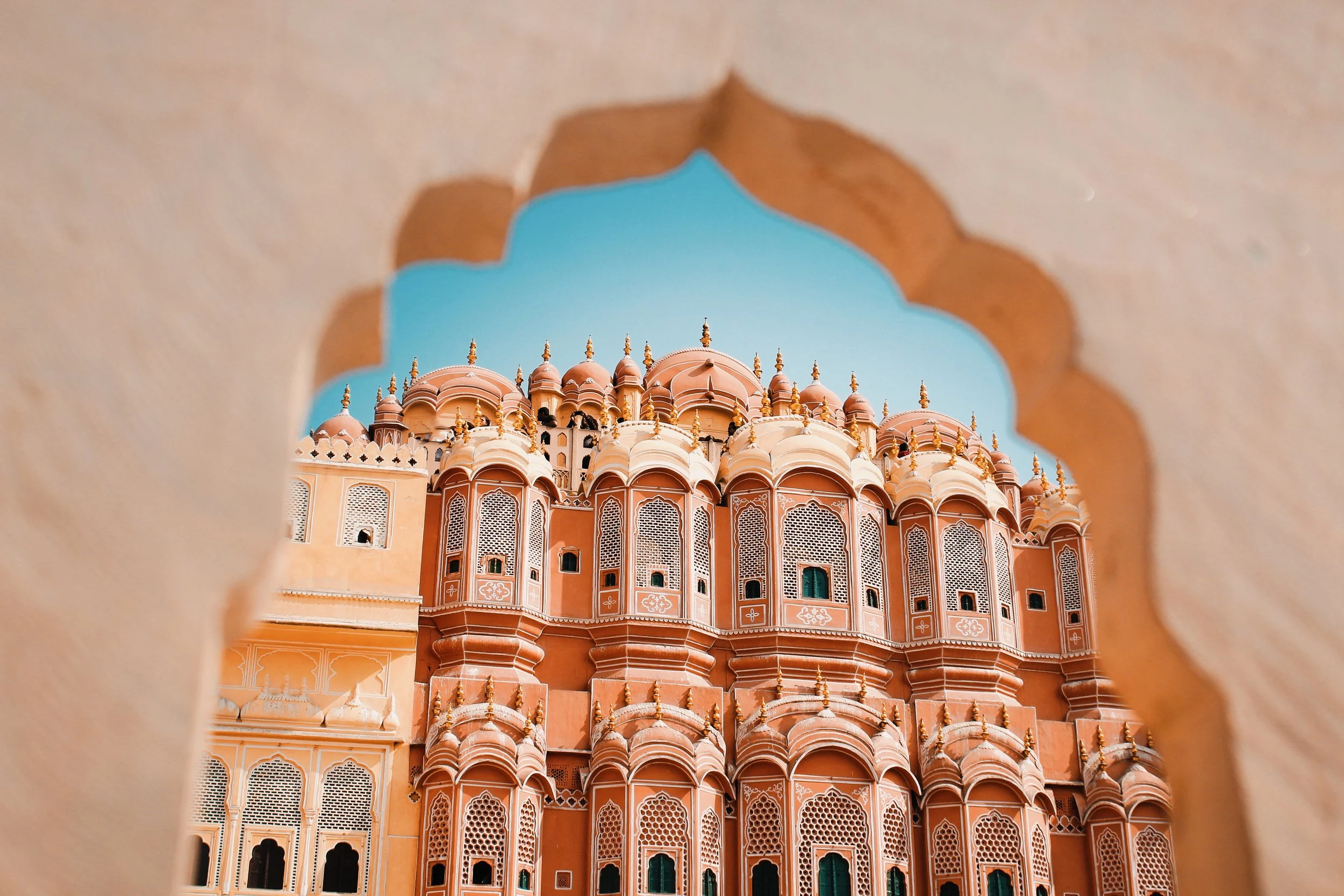 View of an intricate pink and white palace with domes and minarets, seen through a decorative archway with a blue sky in the background.
