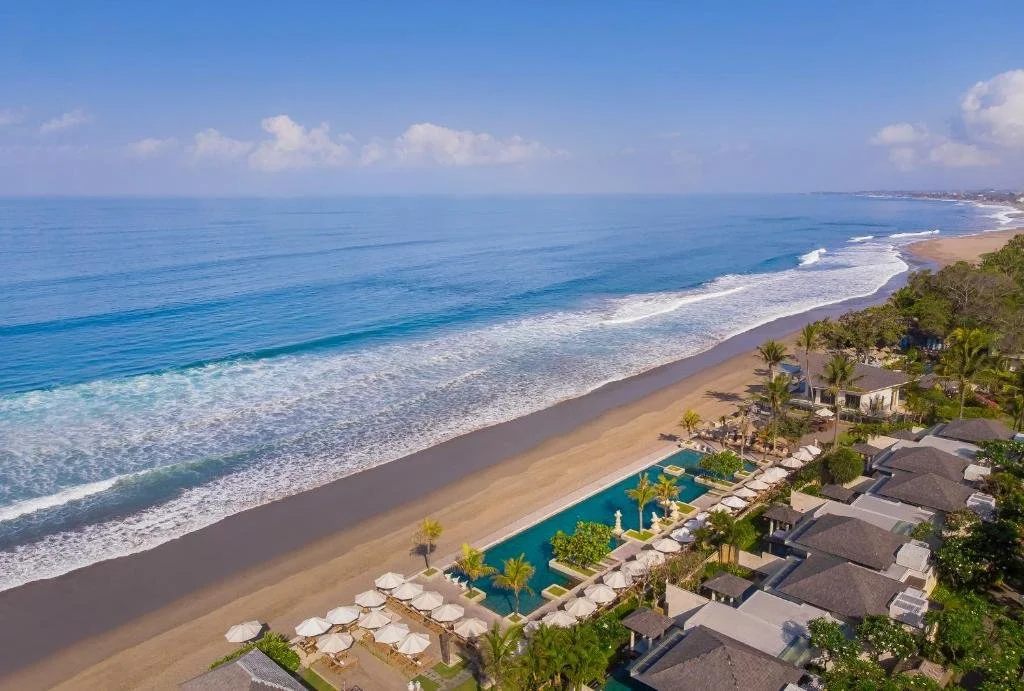 Aerial view of a beachfront resort with a large swimming pool, surrounded by lounge chairs and umbrellas, overlooking a sandy beach and the ocean with waves and a partly cloudy sky.