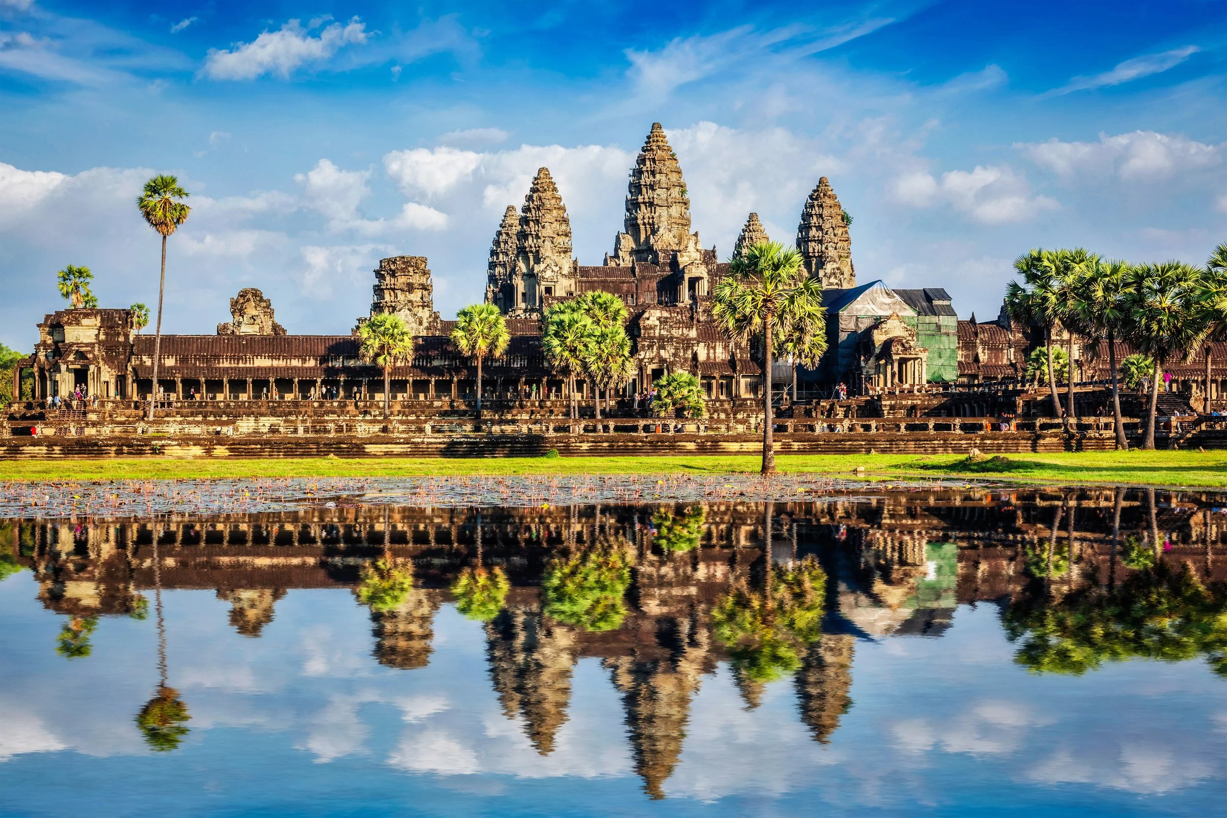 Ancient temple complex with multiple towers, surrounded by lush trees and reflected in a body of water under a partly cloudy sky.