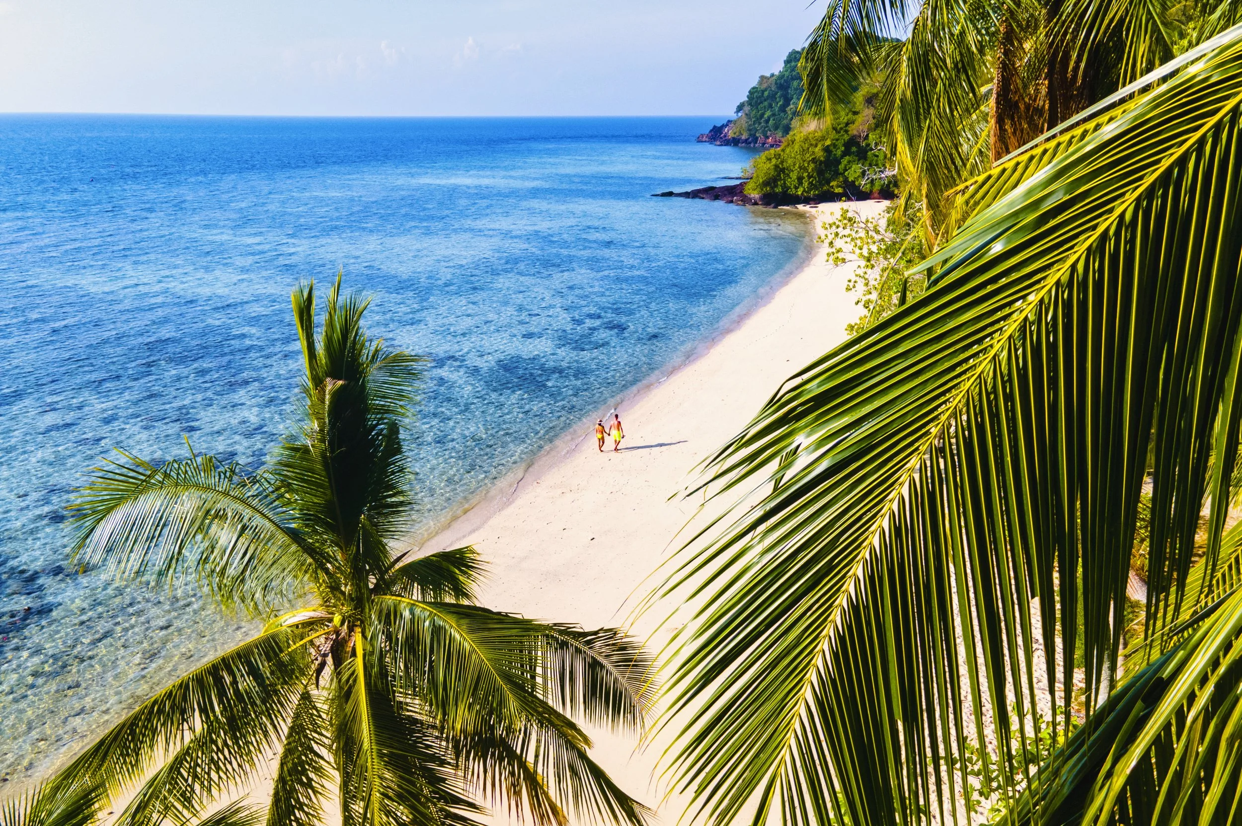 A tropical beach with clear blue water, white sandy shore, and palm trees; two people walking along the beach, with green hills in the background.