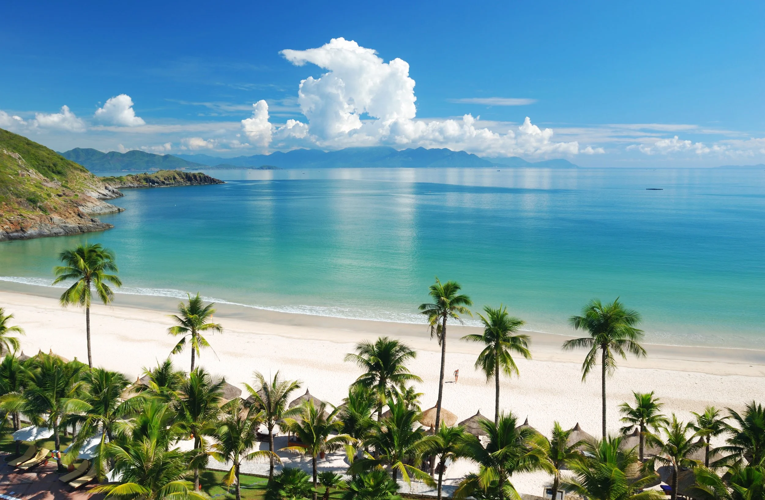 Tropical beach with white sand, numerous palm trees, calm turquoise water, green hills on the left, distant mountains, partly cloudy blue sky.